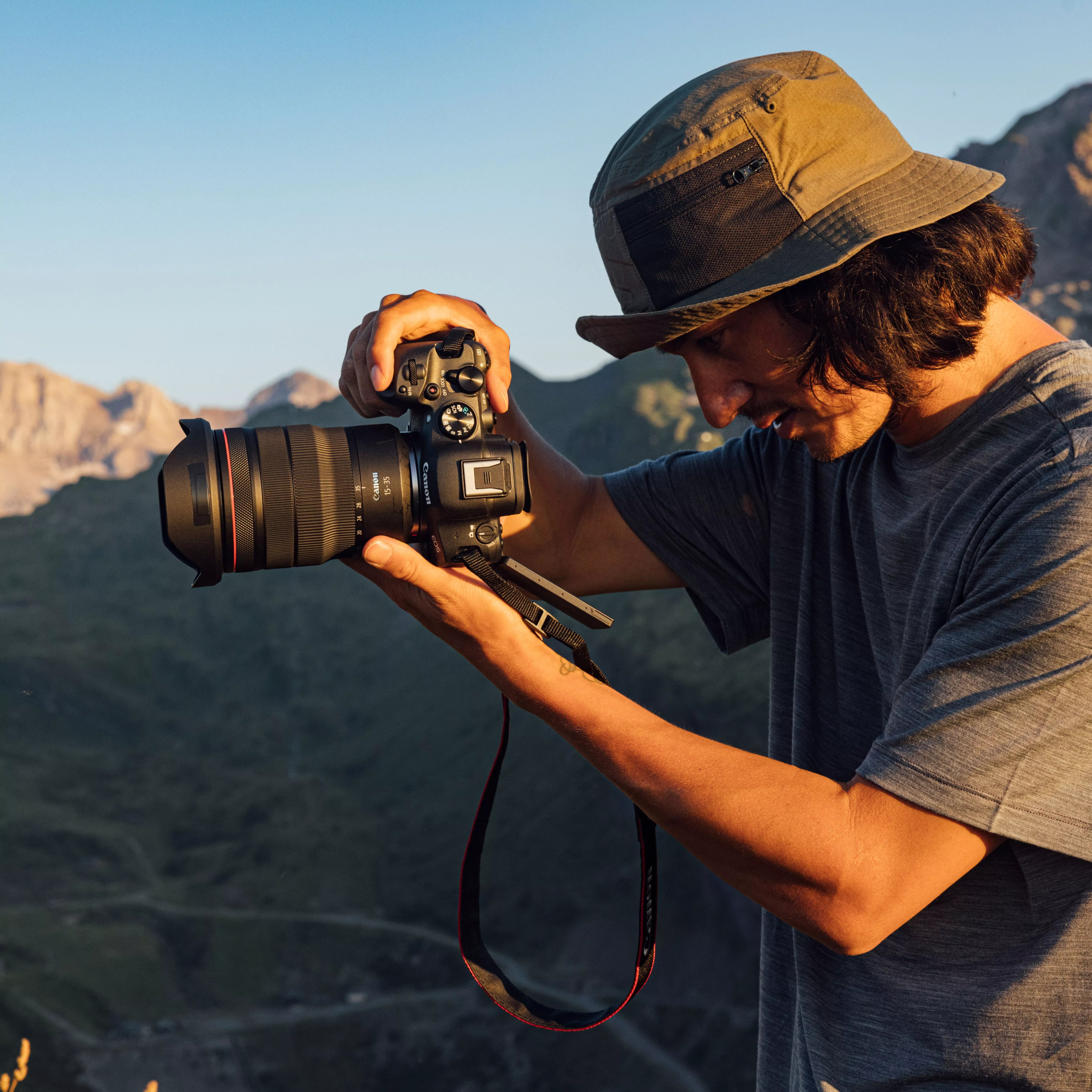 Outdoor photographer with DSLR and telephoto lens capturing mountain landscape at golden hour, wearing sun hat