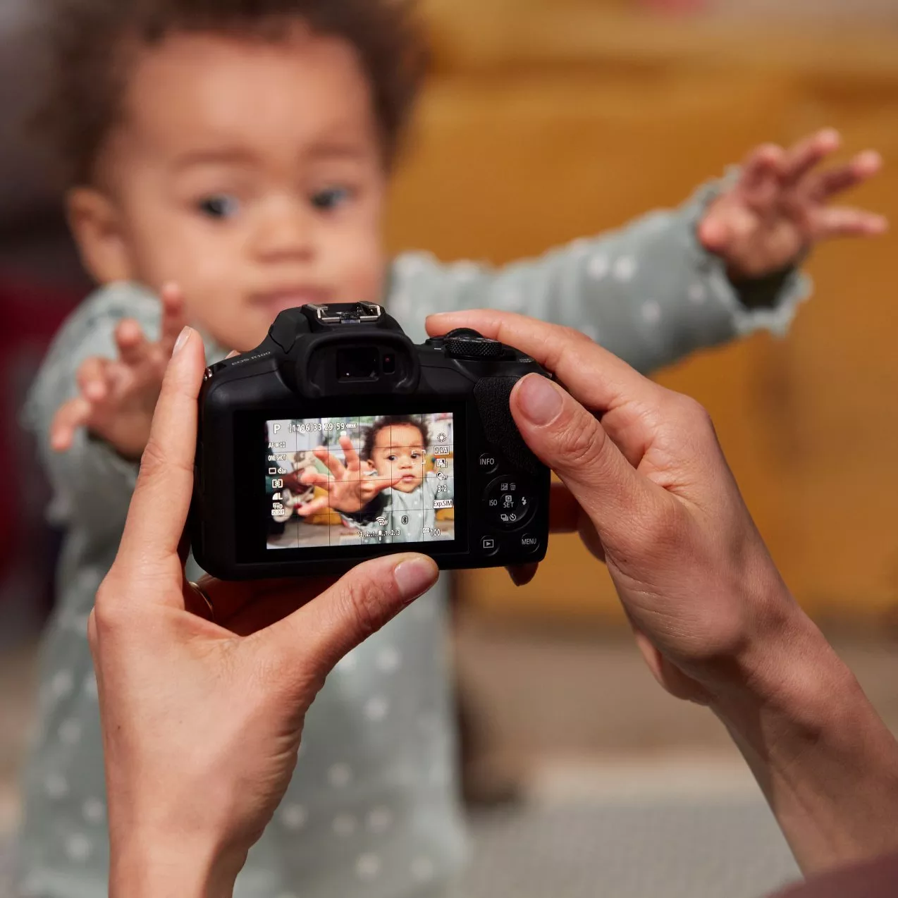 Hands holding a digital camera to photograph a blurred toddler reaching toward the lens, visible on the camera screen.