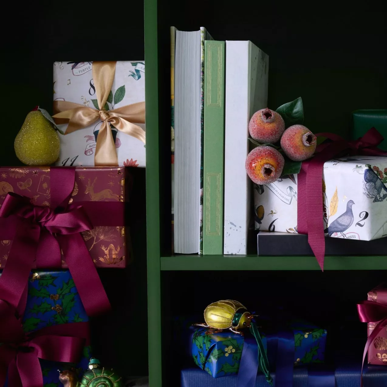 A dark green bookshelf displayed with elegantly wrapped Christmas gifts in floral and patterned paper with satin ribbons, alongside books and festive ornaments like a pear and decorative fruit.