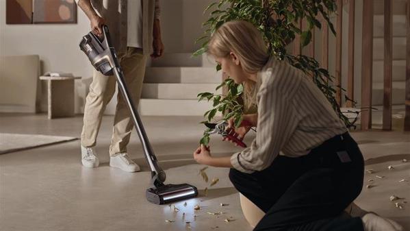 A woman trims a plant while a person vacuums fallen leaves on a modern living room floor.