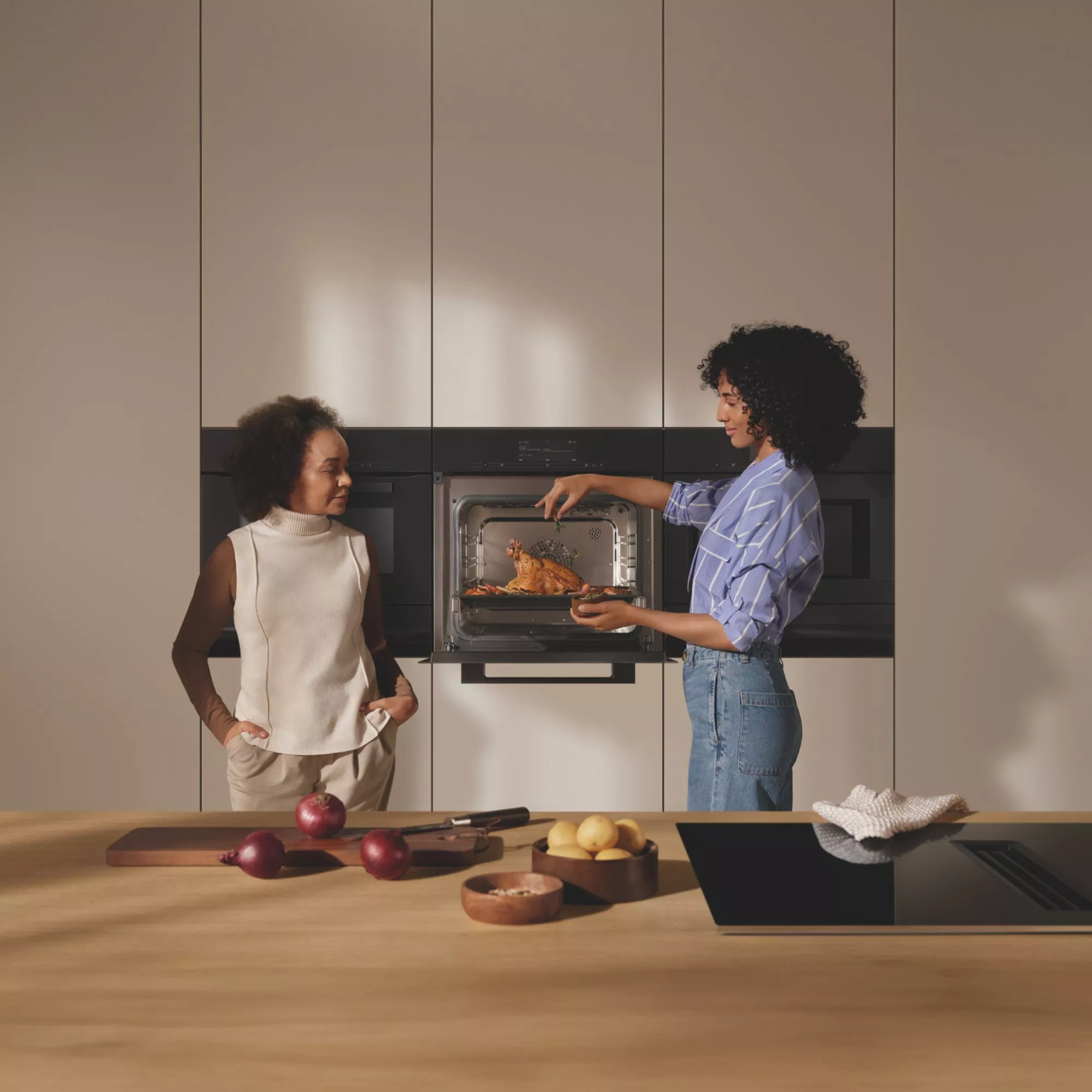 Two people in a modern kitchen, one taking a roast chicken from an oven. Fresh ingredients on the counter.