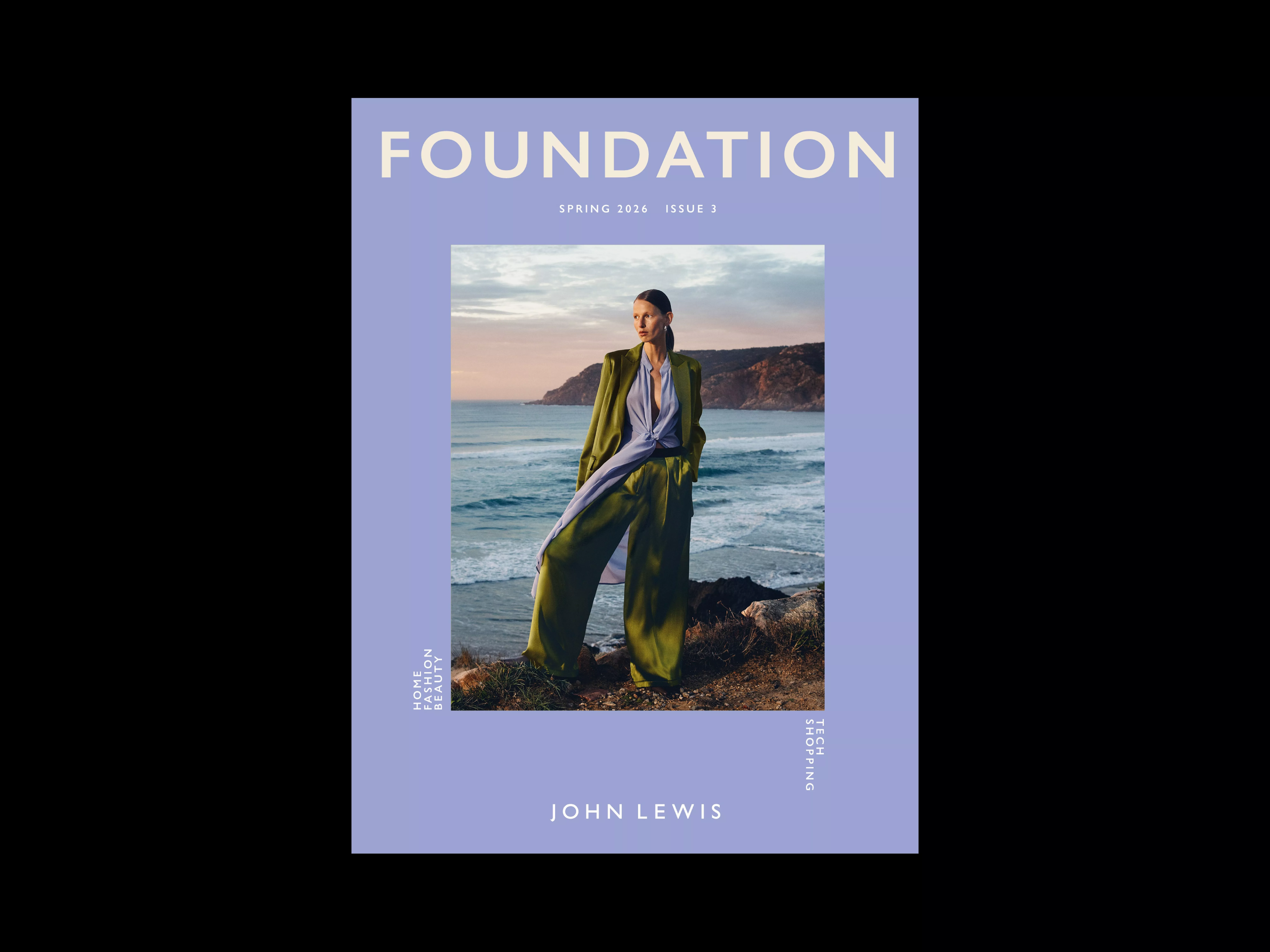 A woman in green trousers and jacket stands by the sea at sunset on the cover of John Lewis' Foundation magazine.