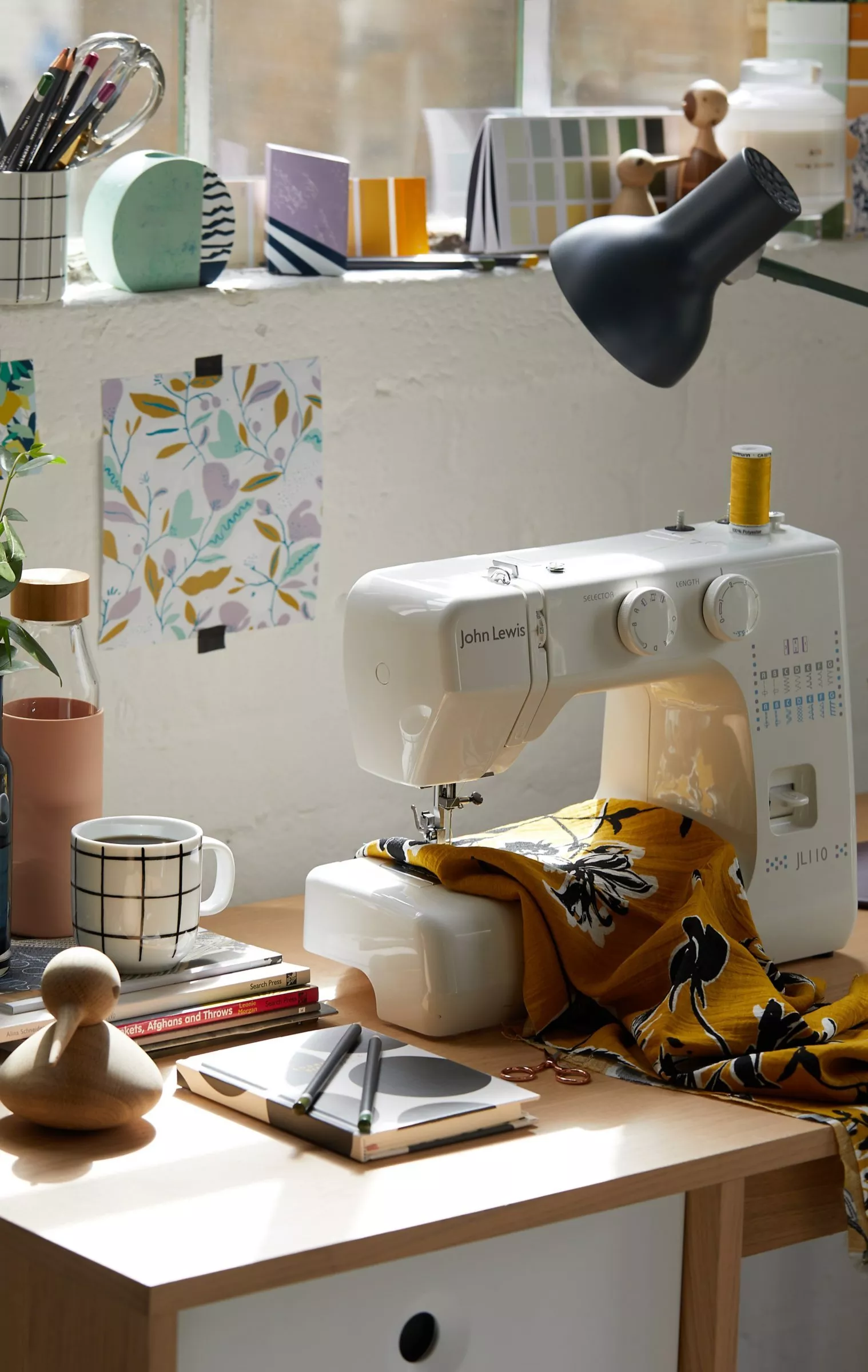 White sewing machine on craft table with patterned fabric, thread spool, lamp and tools in bright studio.