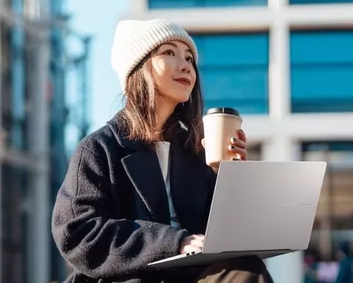 A woman in a beanie sits outdoors, sipping coffee and using a laptop, with a modern building in the background.