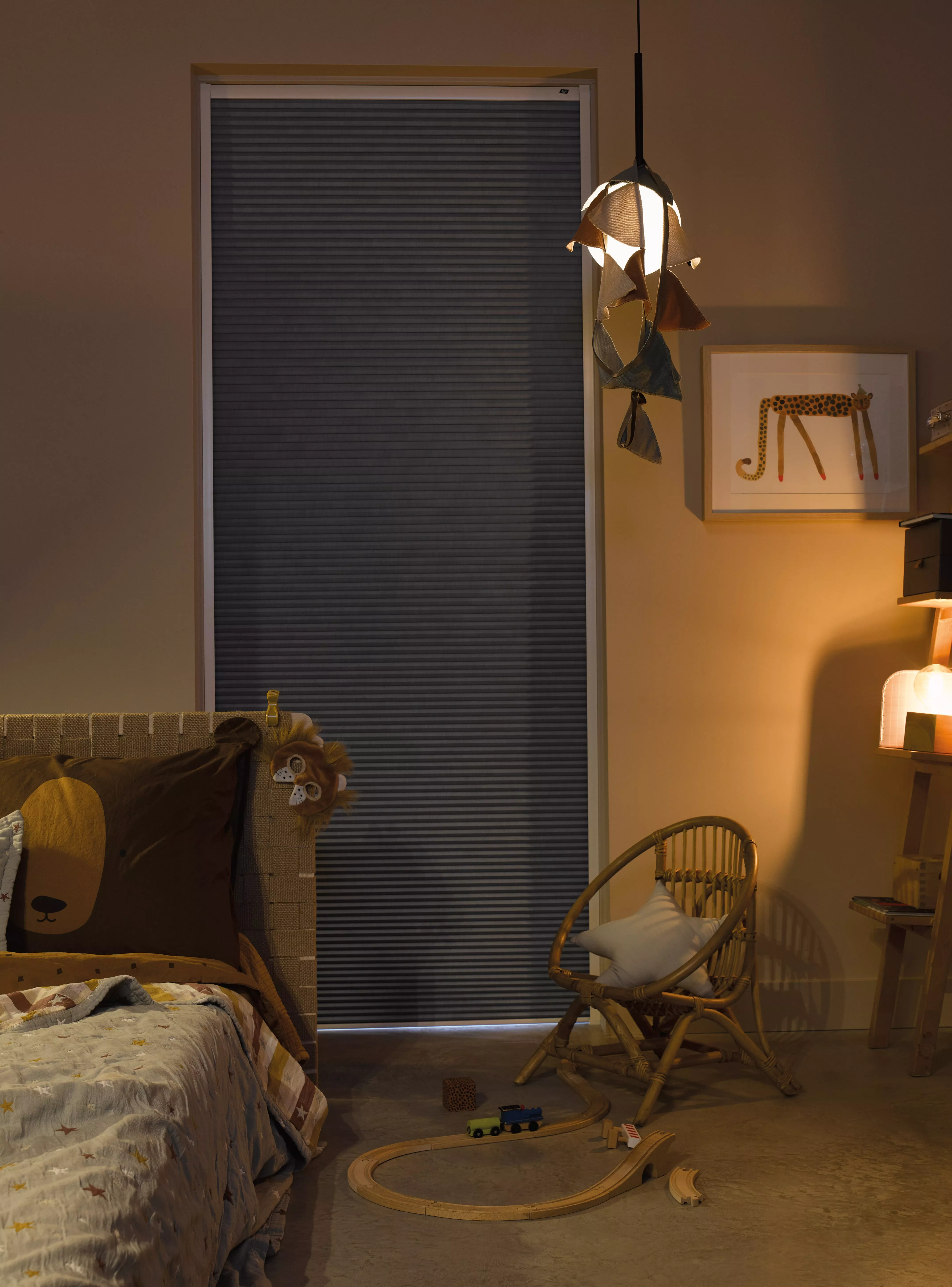 Black pleated blackout blind in a cosy child’s bedroom, with warm pendant light, wicker chair and toy train set