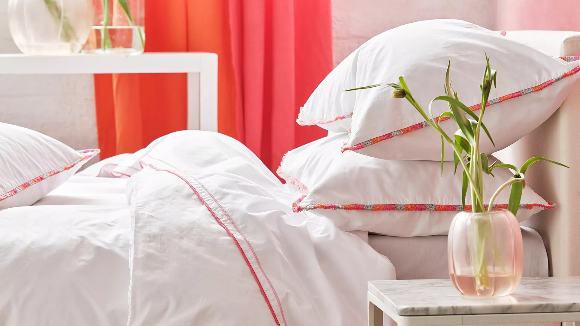 White bedding with pink embroidered trim and stacked plush pillows on a bed, alongside pink sheer curtains and a pink glass vase with green stems on a marble nightstand.