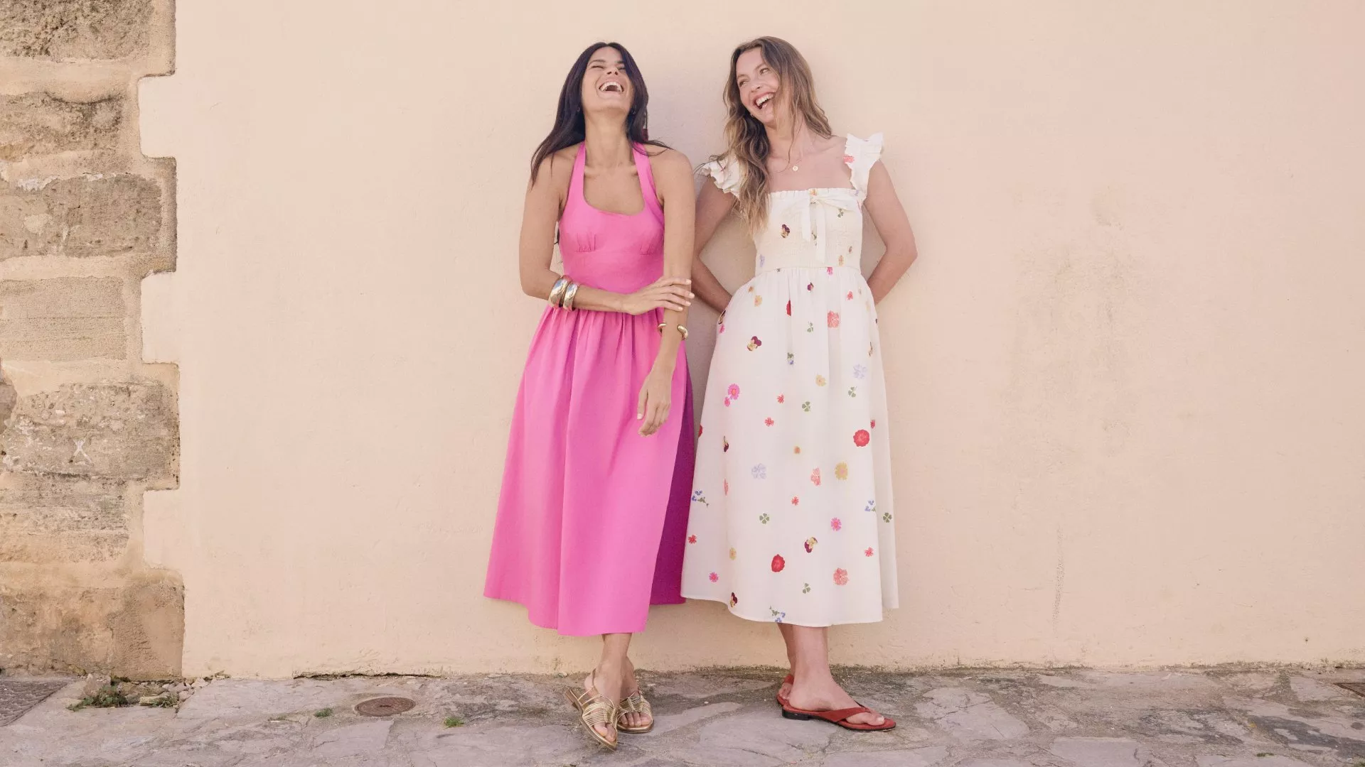 Two women laughing outdoors in summer midi dresses, pink halter dress and white floral dress by a wall