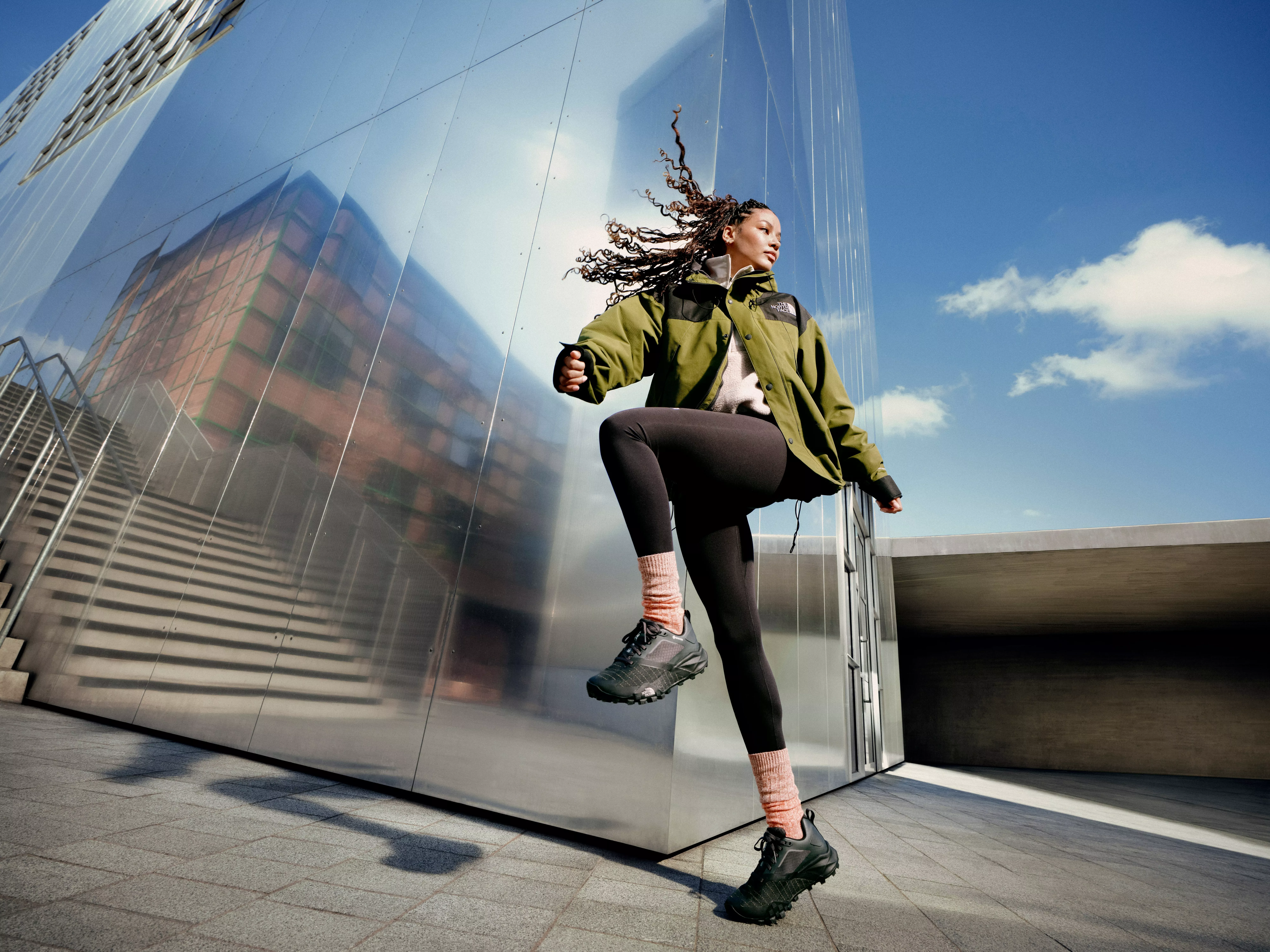 Athlete doing high-knee warm-up outside modern glass building, wearing green jacket, leggings and trail trainers.