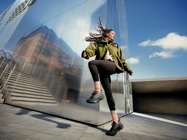 Woman doing high-knee warm-up in outdoor jacket and hiking boots beside modern building, blue sky backdrop