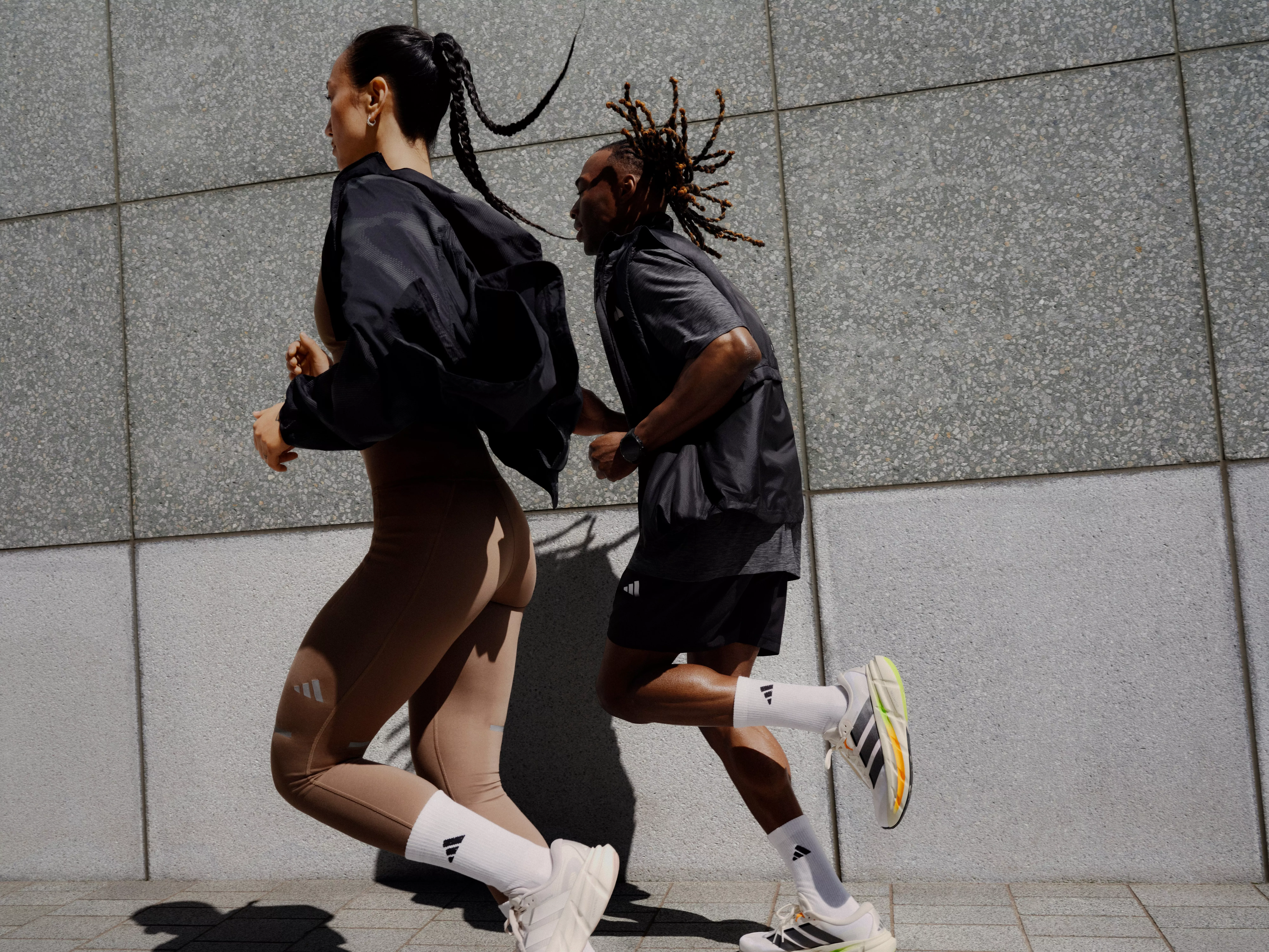 Two runners in modern athletic wear sprinting beside a concrete wall, outdoor fitness training in the city.