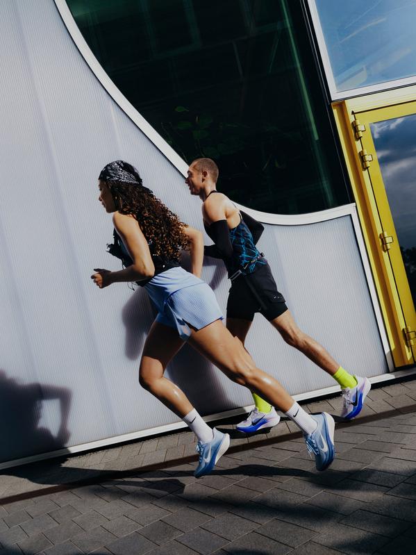Two runners sprinting past a modern building, wearing athletic trainers and sporty outfits in bright daylight.