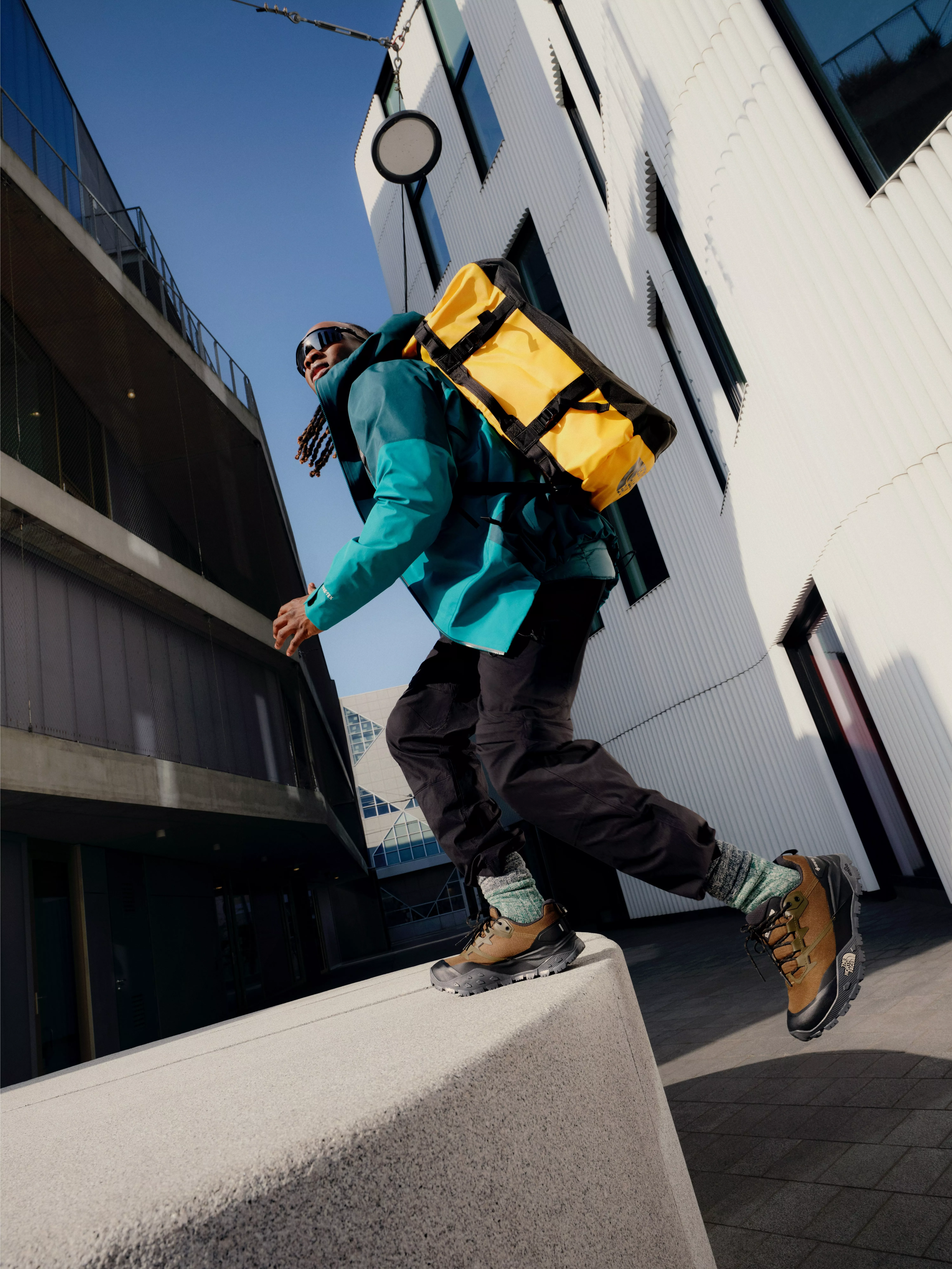 Urban hiker in teal jacket with yellow backpack stepping across concrete ledge between modern buildings outdoors