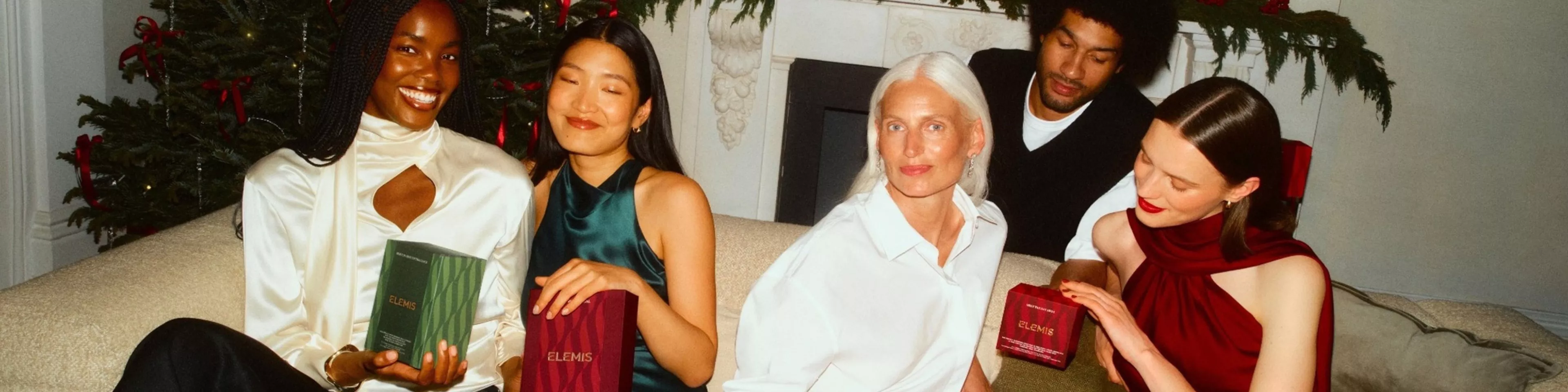 Friends celebrating Christmas in a cosy living room, holding Elemis skincare gift sets by the tree and fireplace.