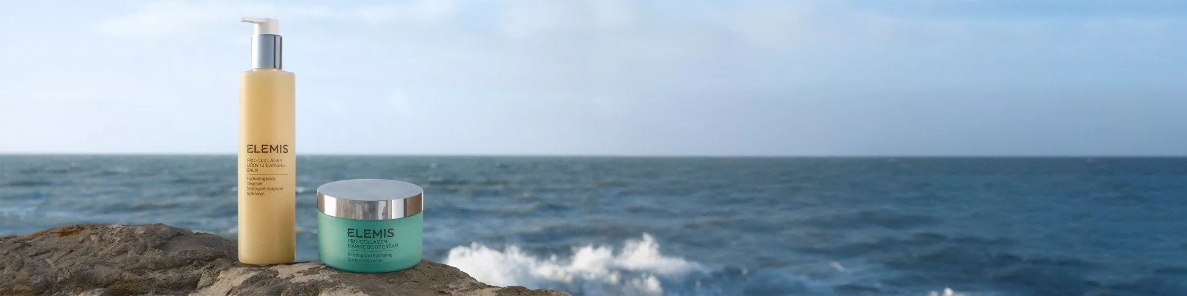 Two Elemis skincare products, a cleansing balm and a body cream, are placed on a rock by the sea.