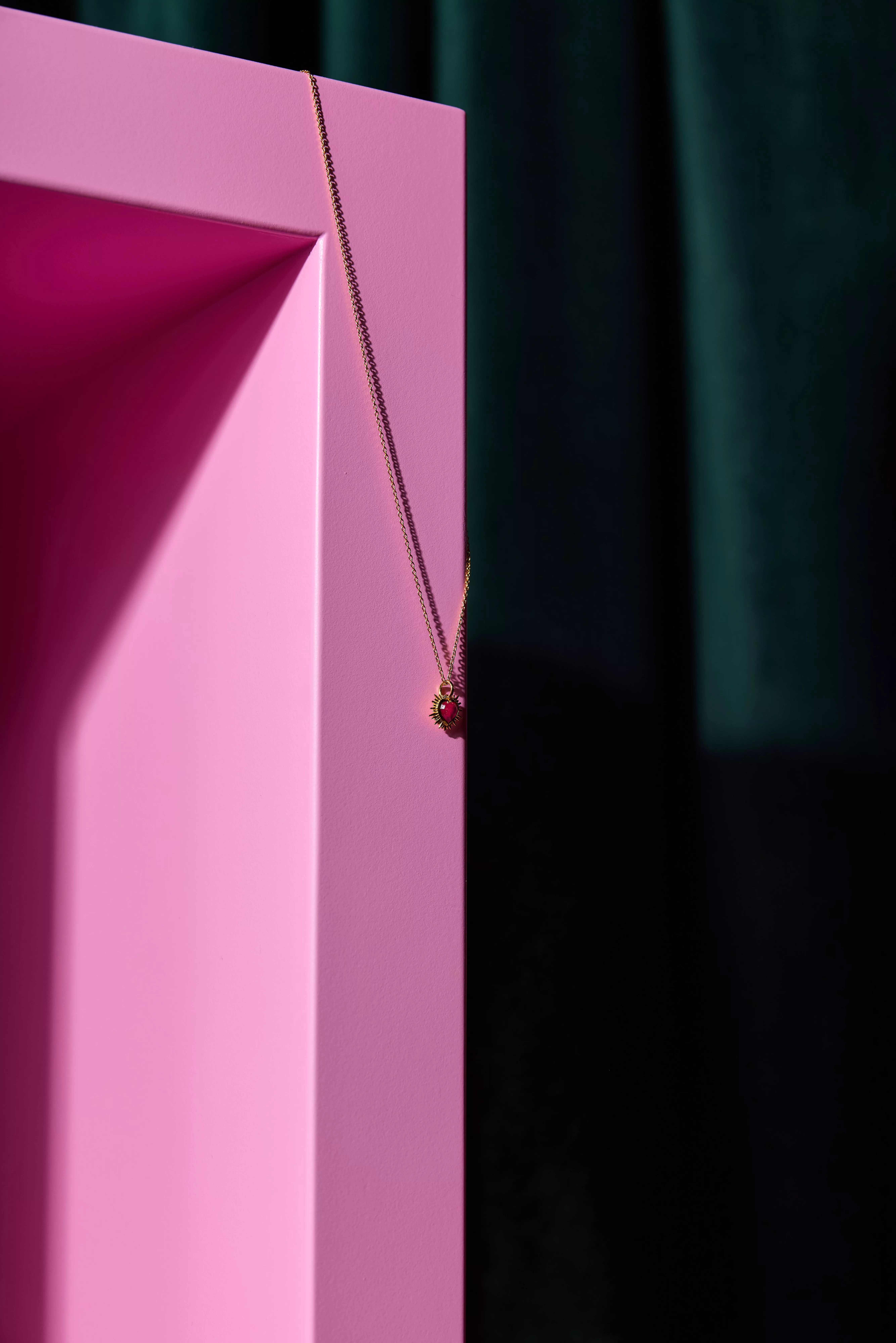 Gold chain necklace with small heart pendant hanging on a pink display block, moody studio lighting background