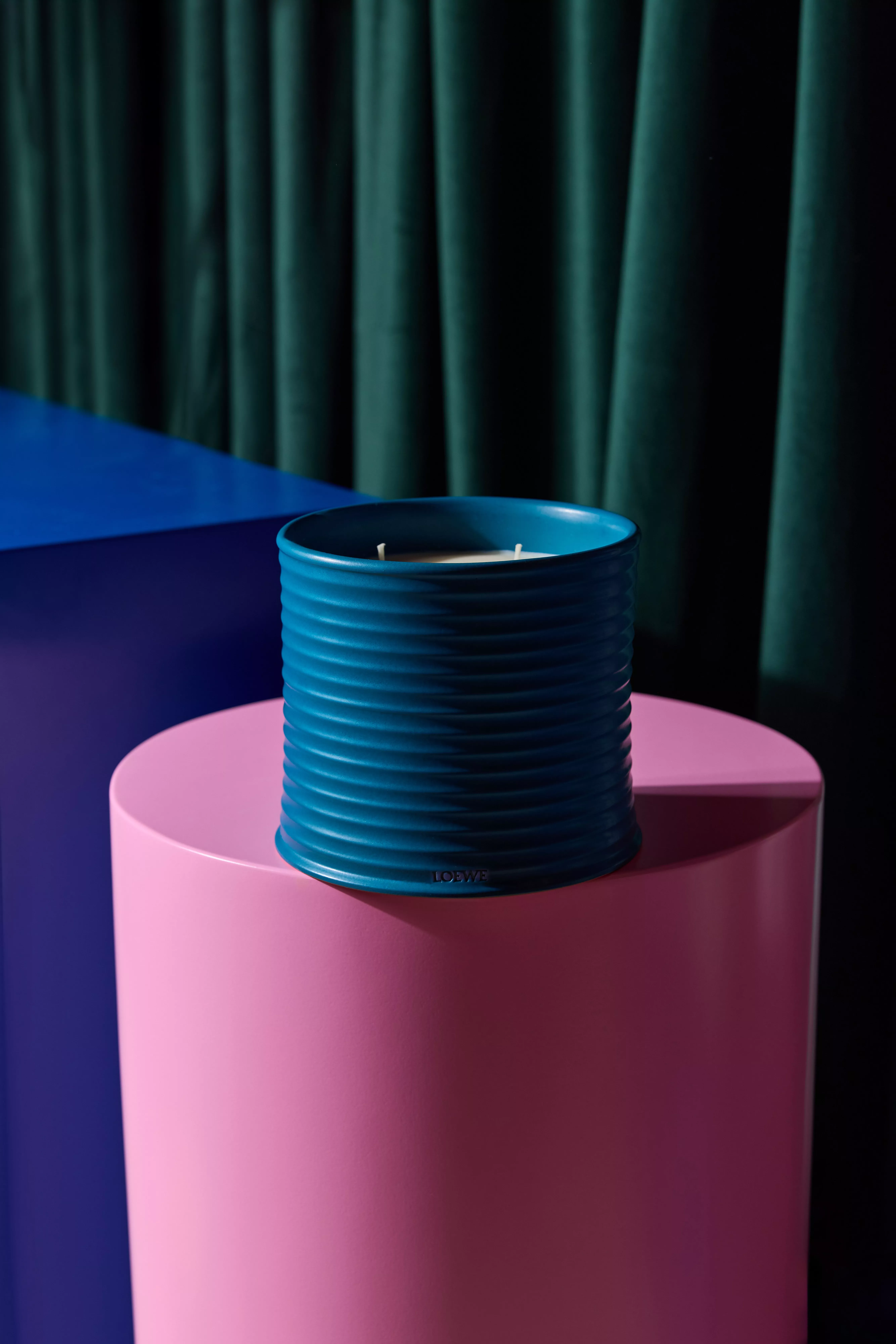 Blue ribbed ceramic scented candle on a pink pedestal, modern home décor product photo with teal curtain backdrop