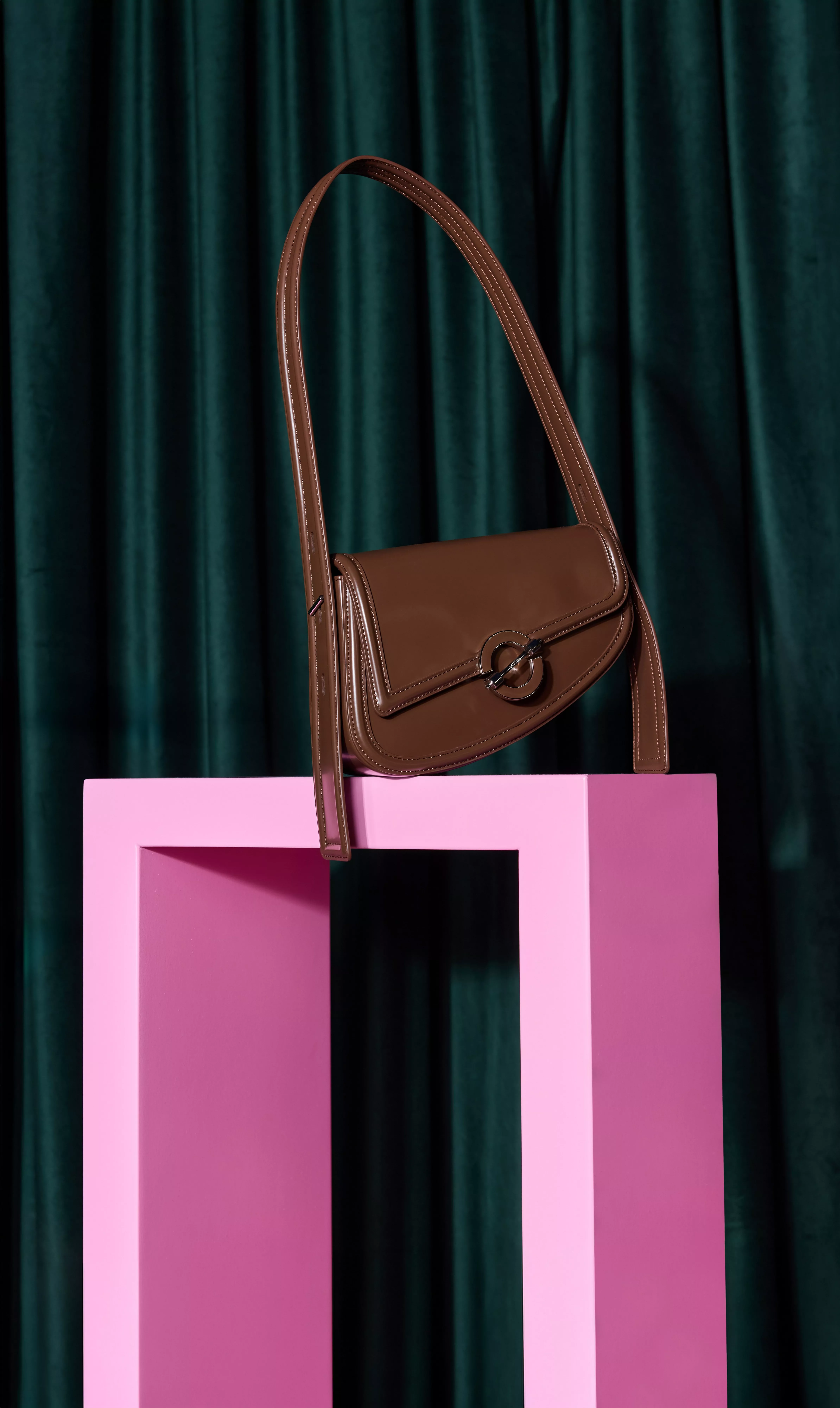 Brown leather shoulder bag on pink plinth against teal curtains, minimalist luxury fashion product photo