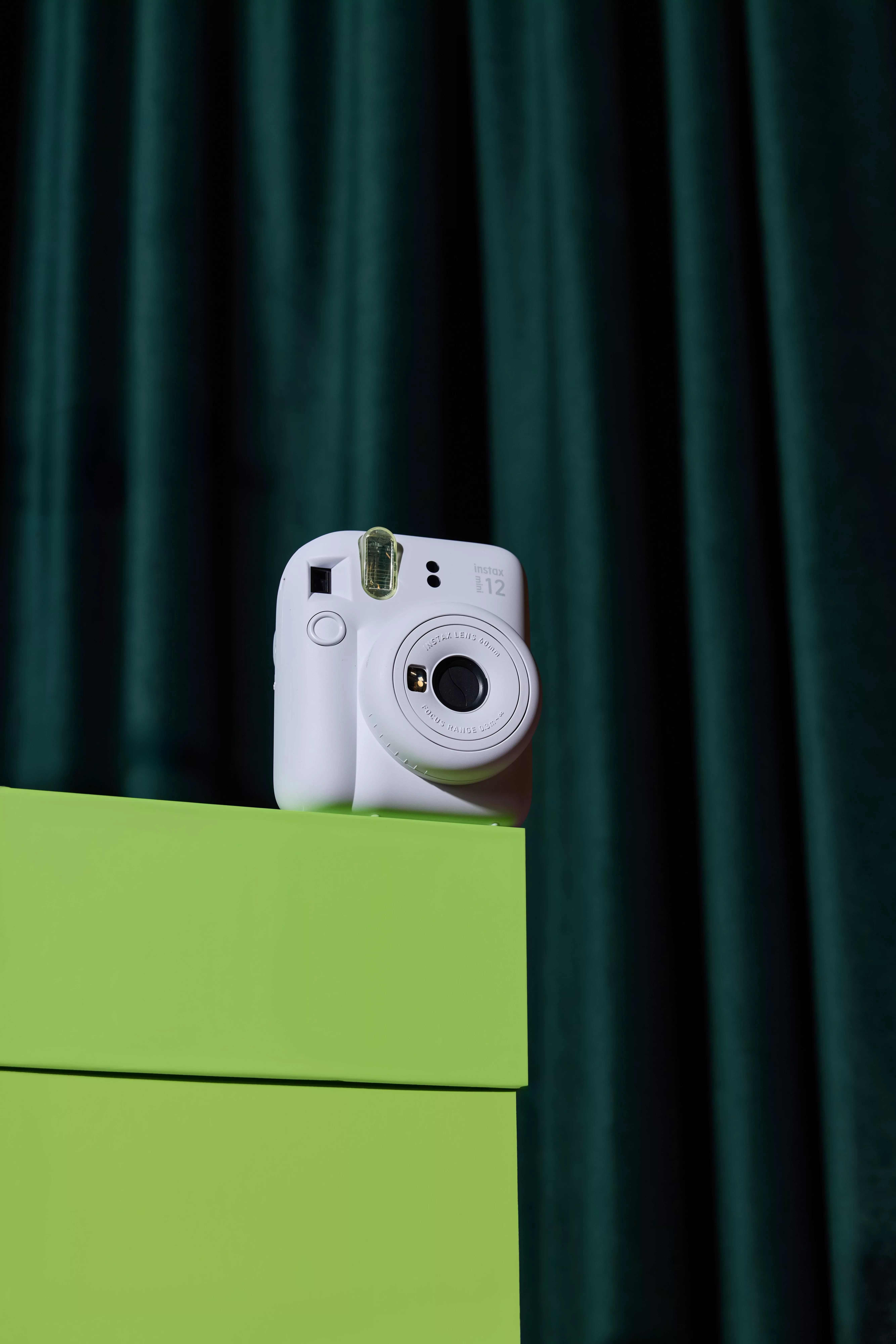 White Instax Mini 12 instant camera on a lime green box against a dark teal curtain backdrop, product photo