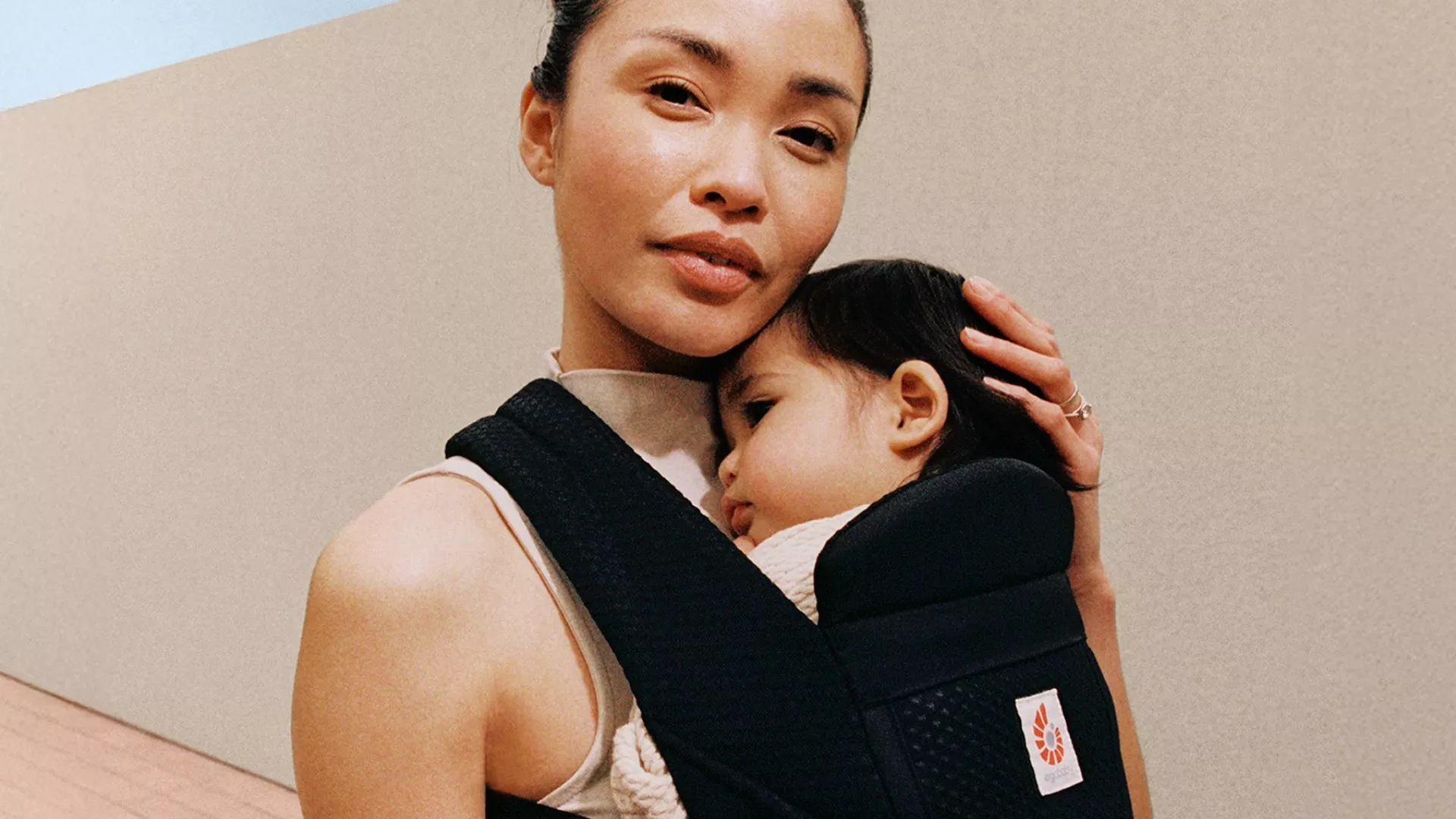 A mother holding her sleeping baby securely in a black Ergobaby carrier while standing near a modern wall.
