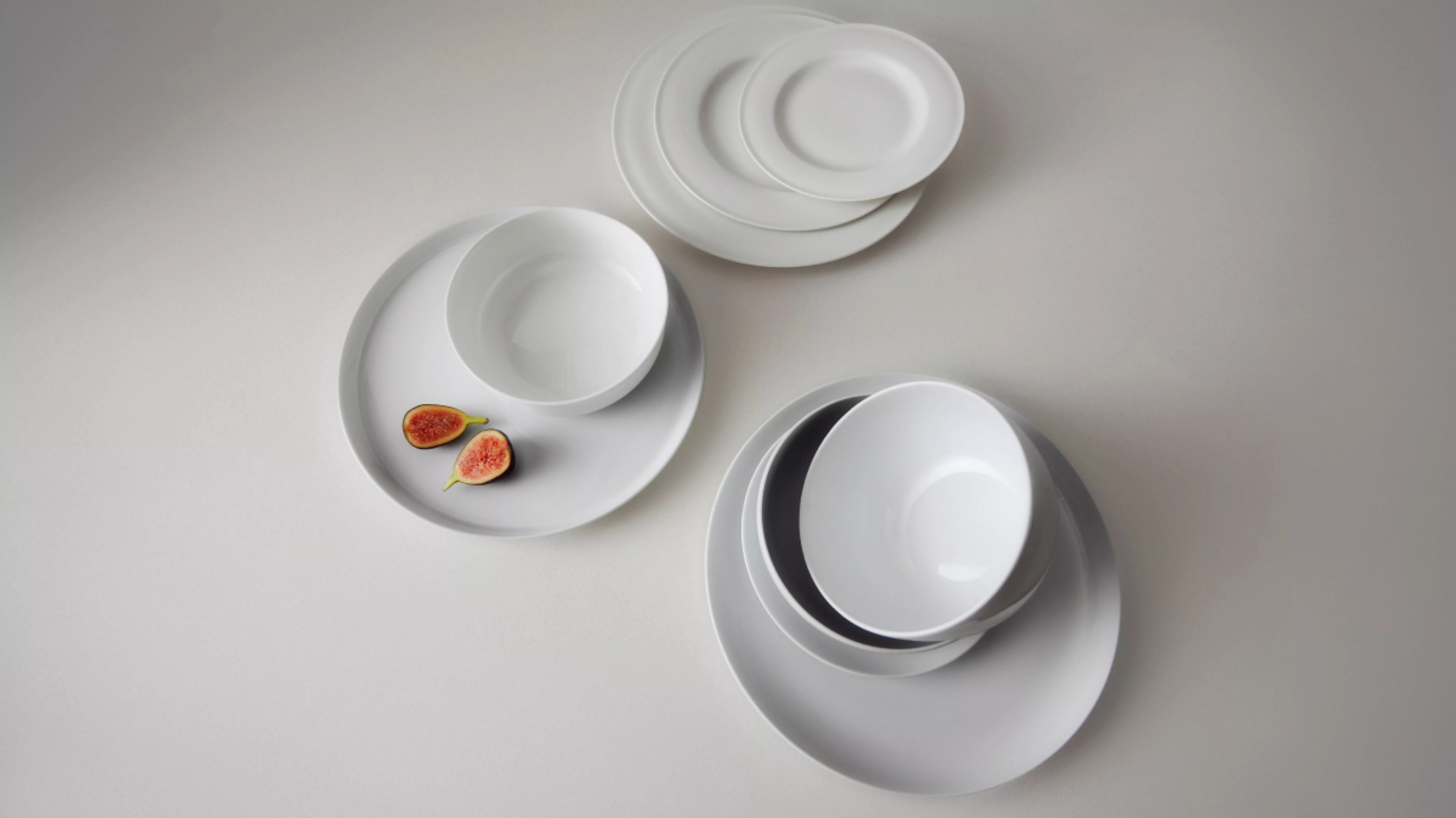Minimalist white porcelain dinnerware set with stacked plates and bowls on grey surface, figs on side plate