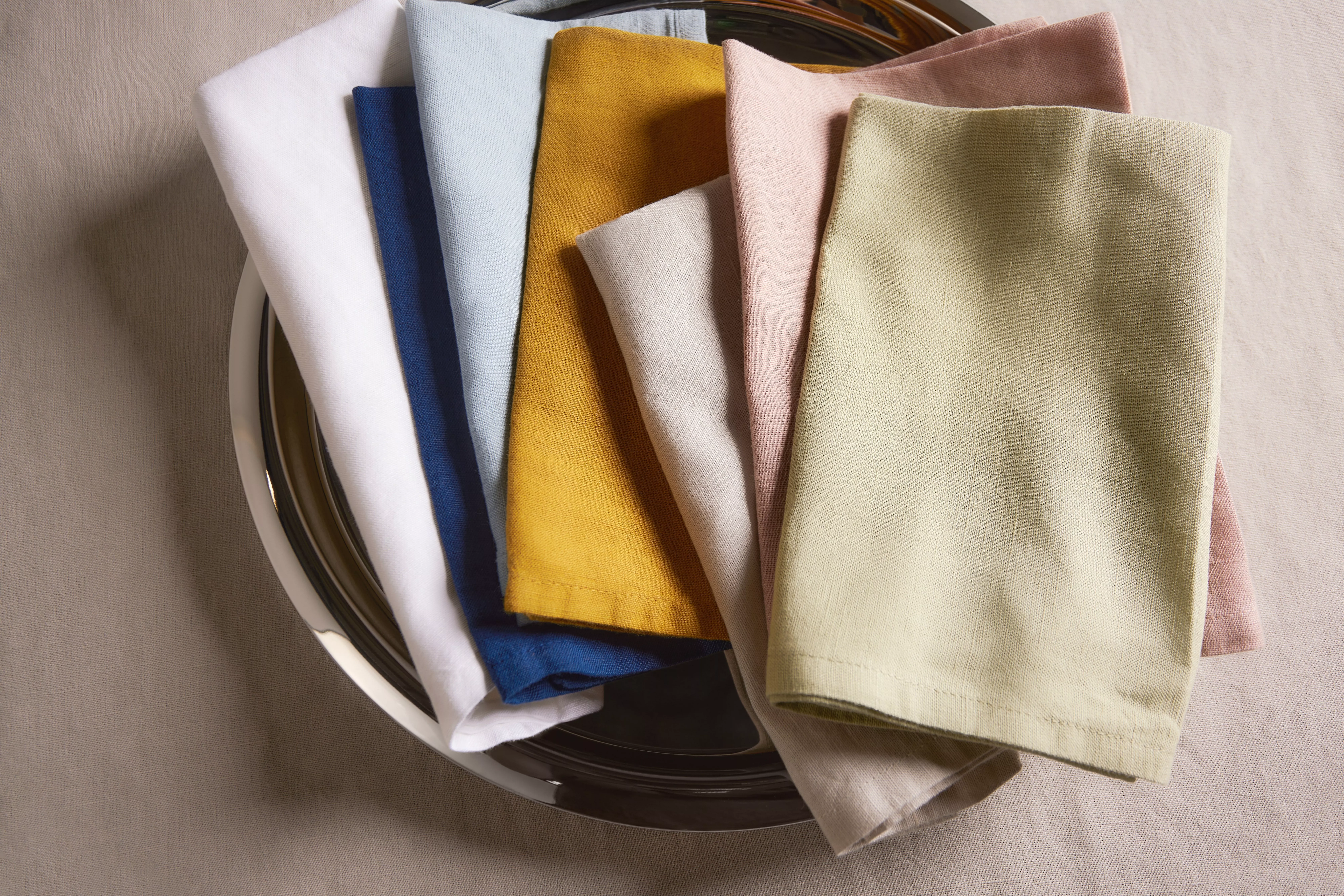 Folded linen napkins in sage green and beige on a white plate, styled on a blue tablecloth for dining decor