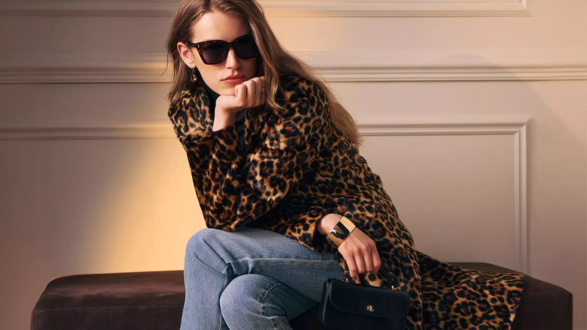 Stylish woman in leopard print coat and sunglasses, seated indoors with jeans and black handbag, fashion portrait