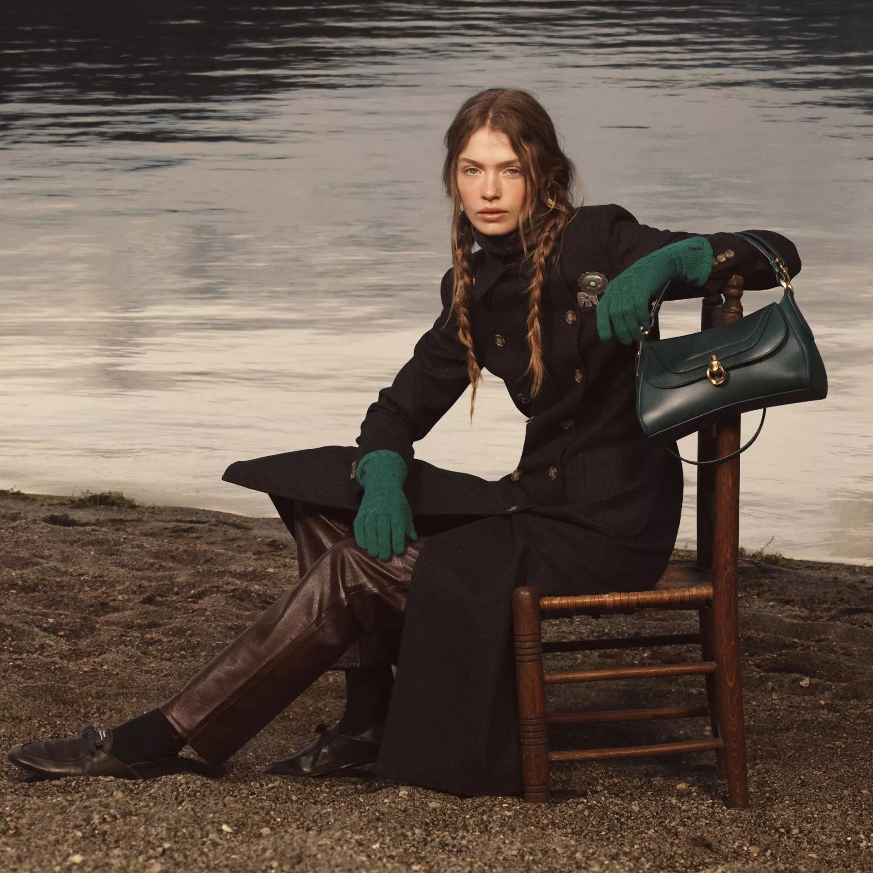 Fashion model by lakeside wearing black coat and green gloves, holding teal leather handbag on wooden chair
