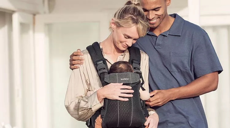 A couple smiling at their baby in a black carrier, enjoying a tender family moment indoors.