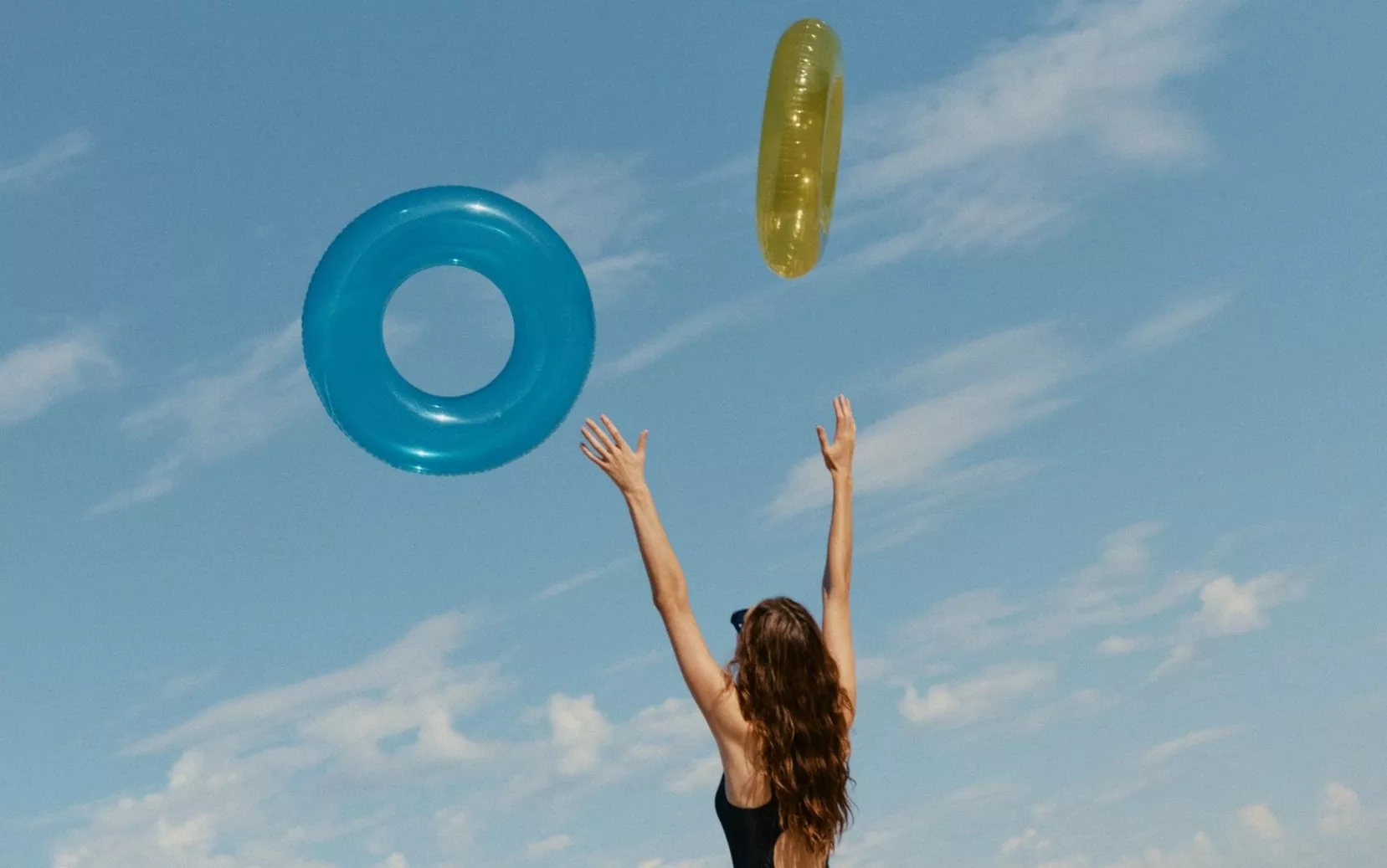 Woman in black swimsuit on sunny beach tossing inflatable rings into blue sky, summer holiday vibe