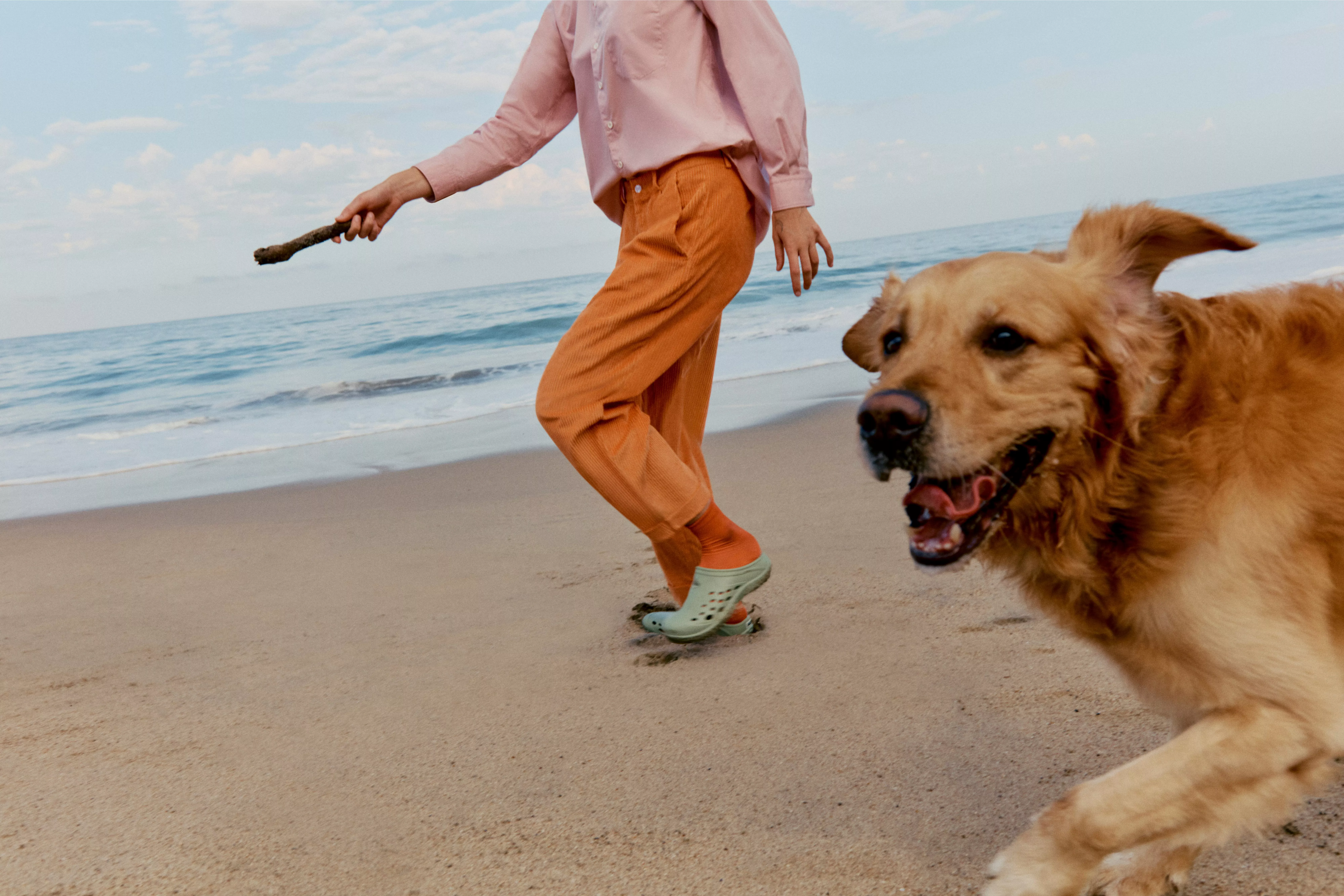 Golden retriever running on sandy beach beside person holding a stick, with calm sea and blue sky backdrop