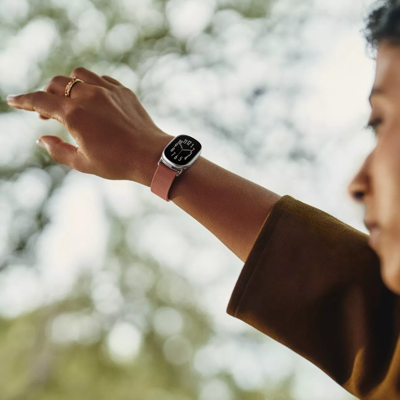 Close-up of a person wearing a silver smartwatch with an analog clock face and a pinkish-brown strap, raised outdoors against a tree background.