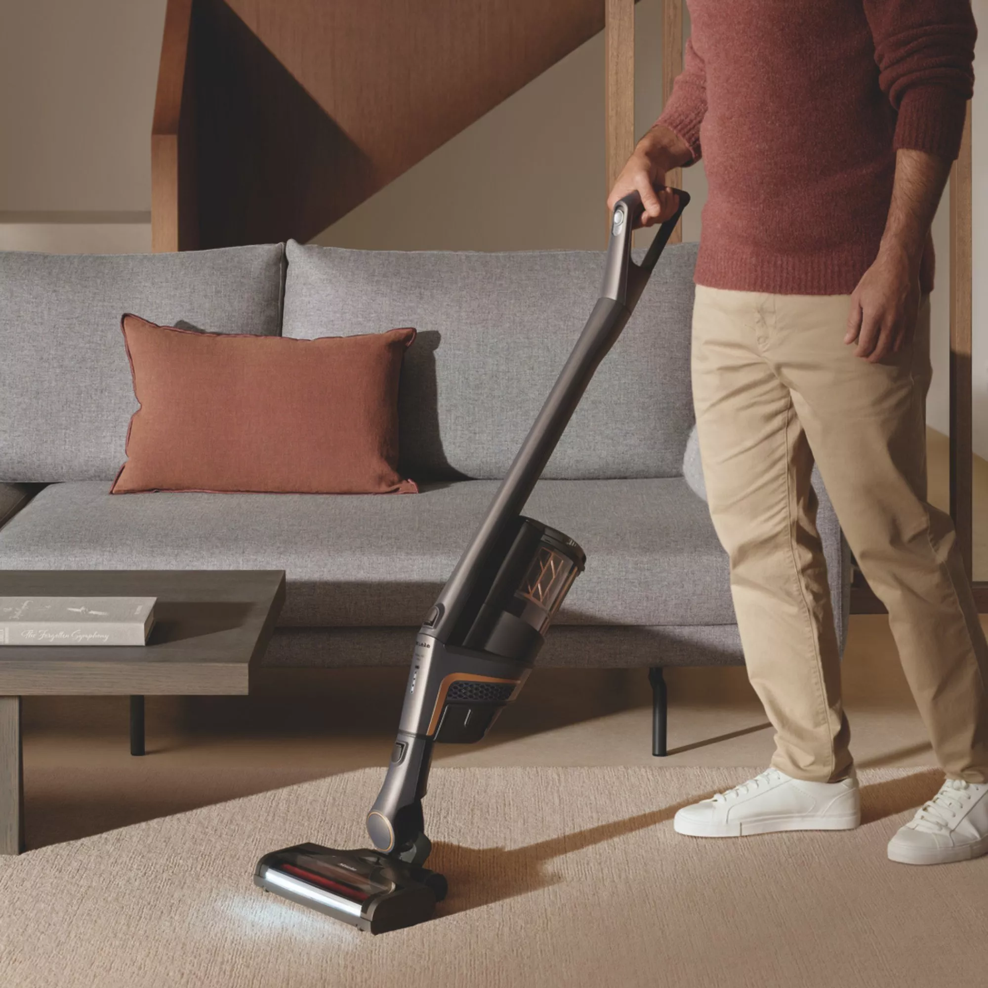 Person vacuuming a beige carpet in a modern living room with a cordless stick vacuum cleaner and grey sofa in background