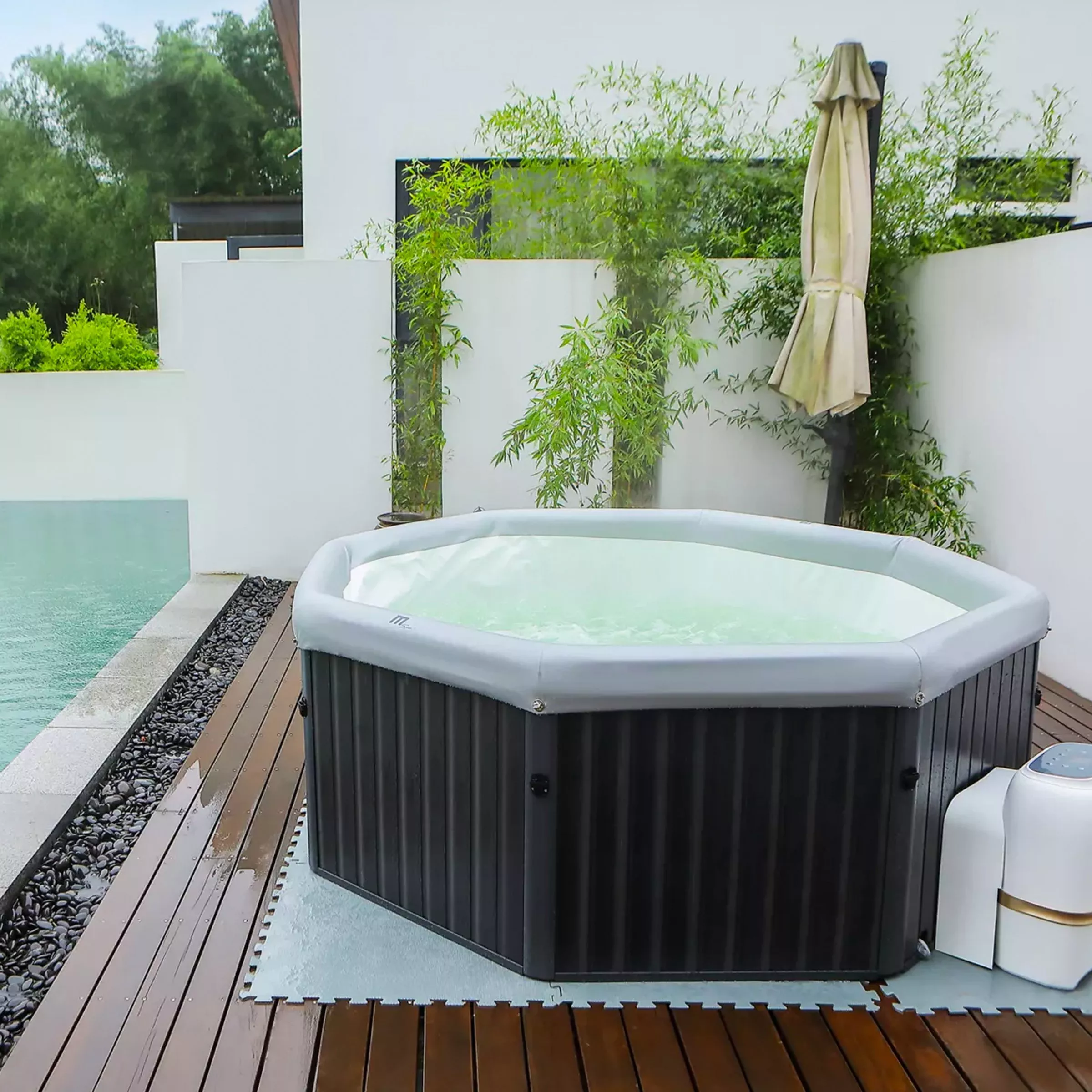 Outdoor inflatable hot tub on wooden decking by pool, with bamboo plants and closed patio umbrella backdrop