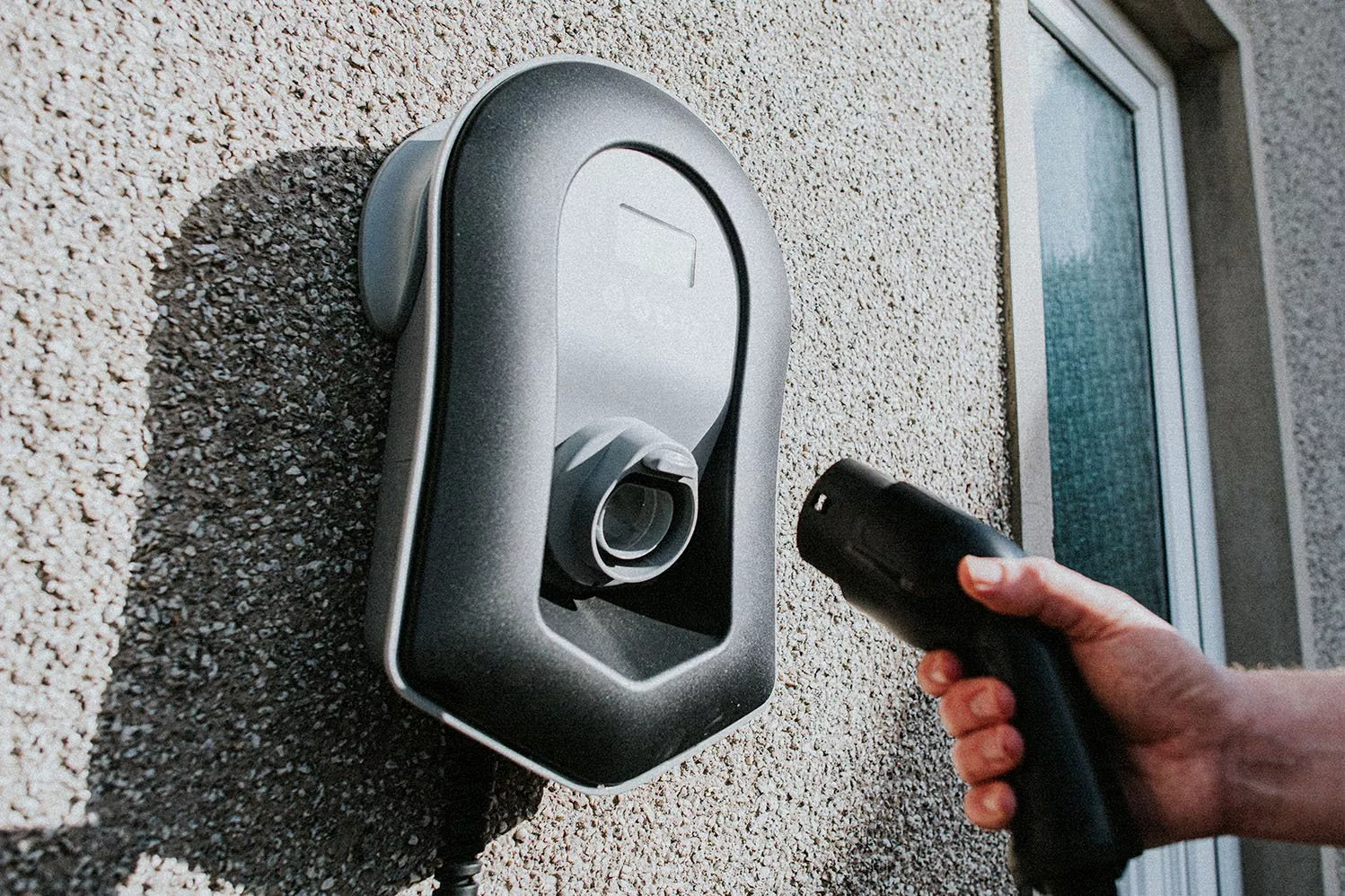 Hand plugging electric car charging cable into wall-mounted home EV charger, close-up by exterior window