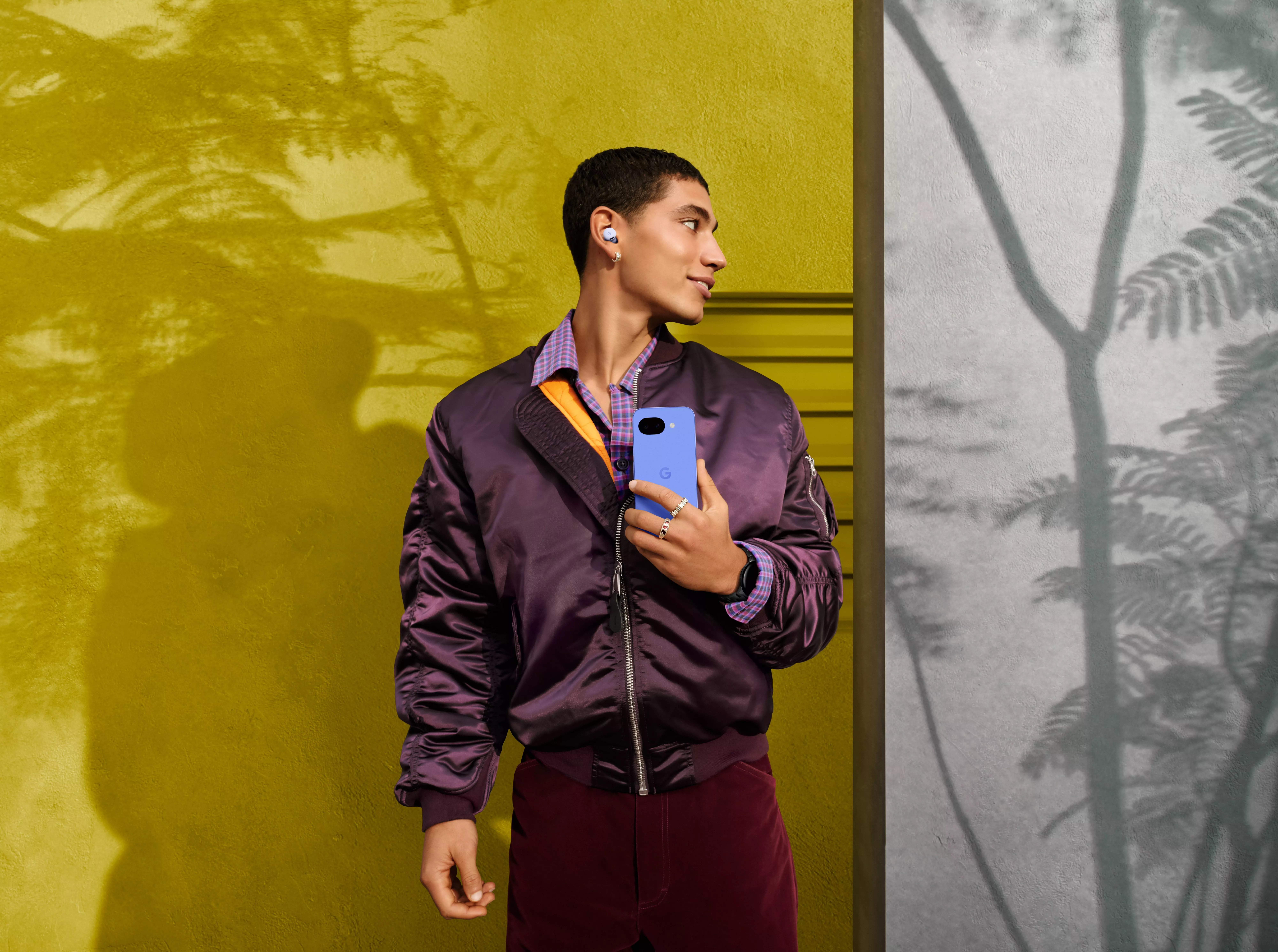 Person holding a blue Google Pixel smartphone and wearing wireless earbuds, standing by a yellow wall outdoors
