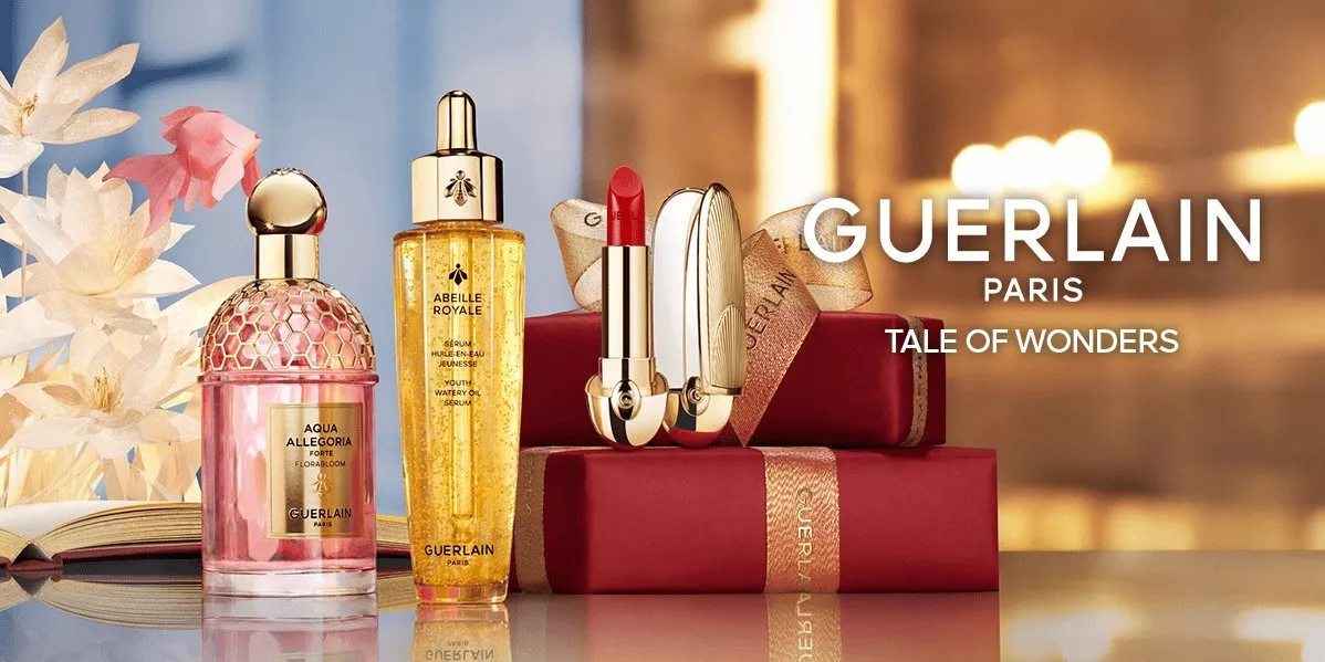 Showing Guerlain oil serum, red lipstick and a perfume bottle surrounded by flowers and a red gift box.