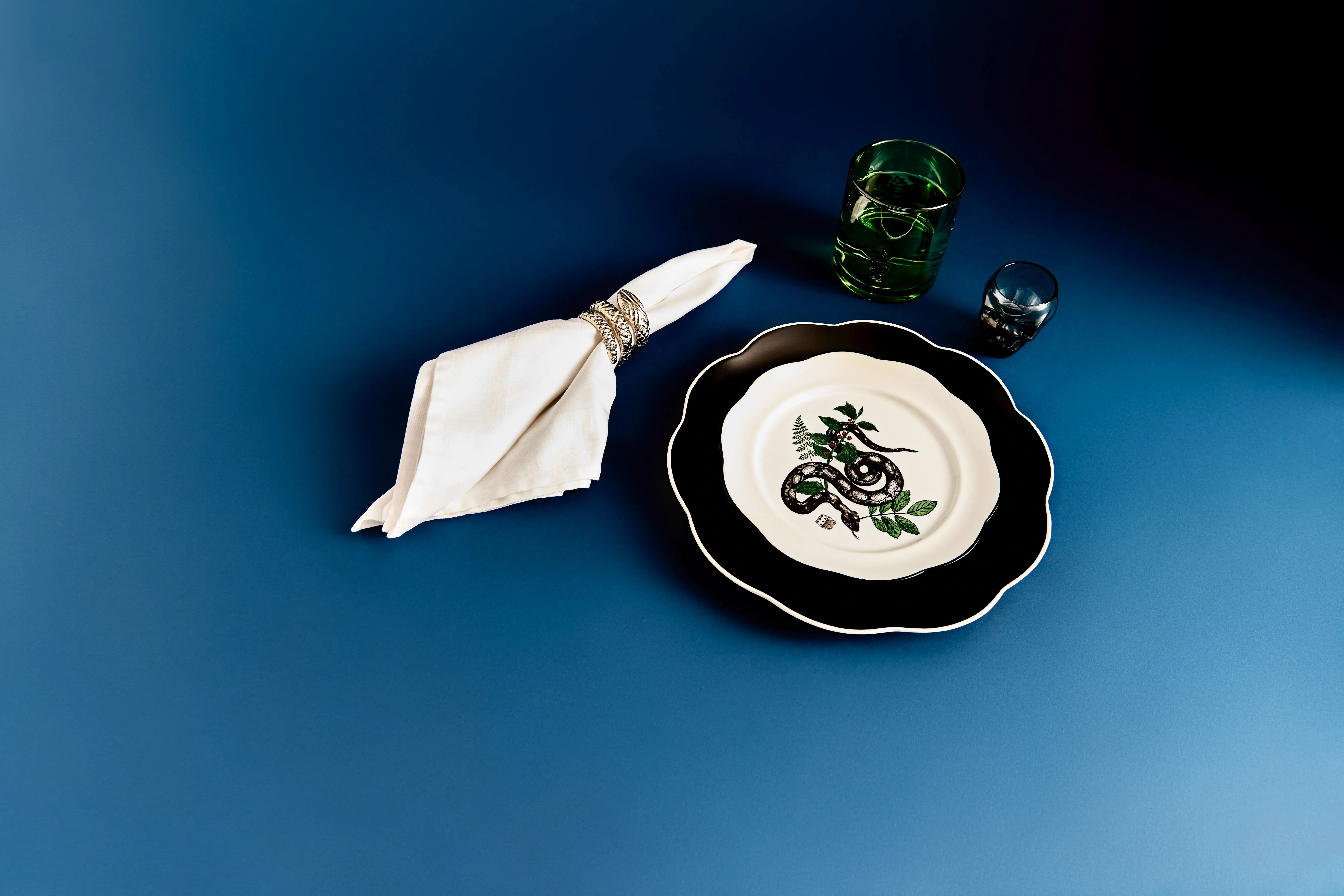 Stylish table setting on blue background with snake motif plate, white napkin ring, and green glass tumbler