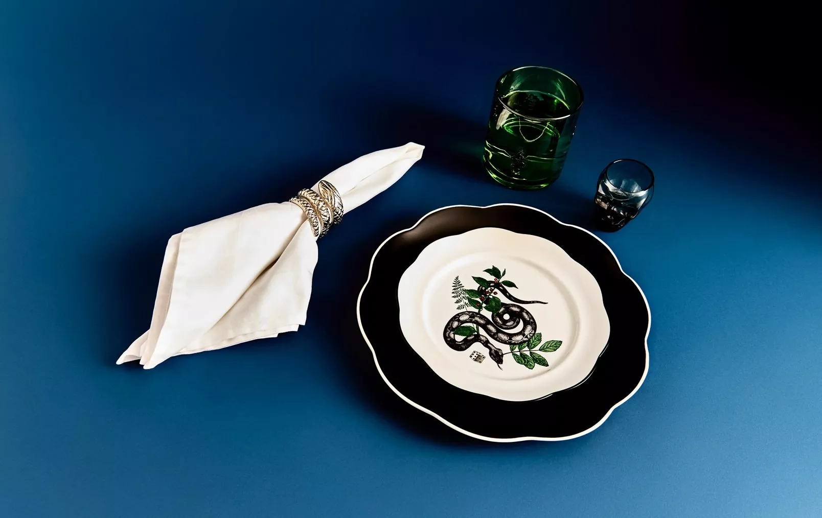 Stylish table setting with snake motif dinner plate, white napkin ring, and green glassware on blue background
