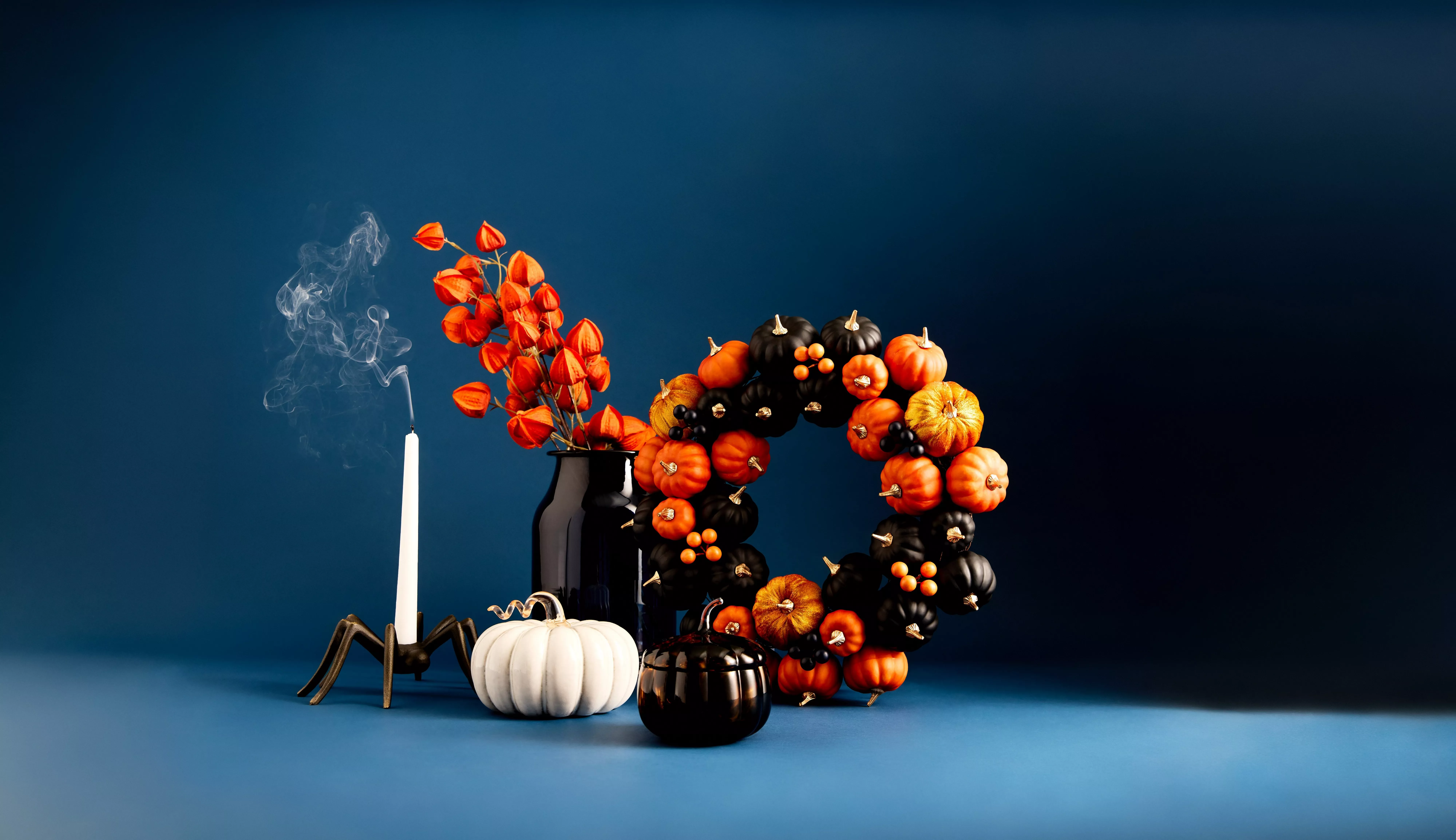 Halloween still life with pumpkins, black wreath, smoky candle, spider holder and orange flowers on blue backdrop