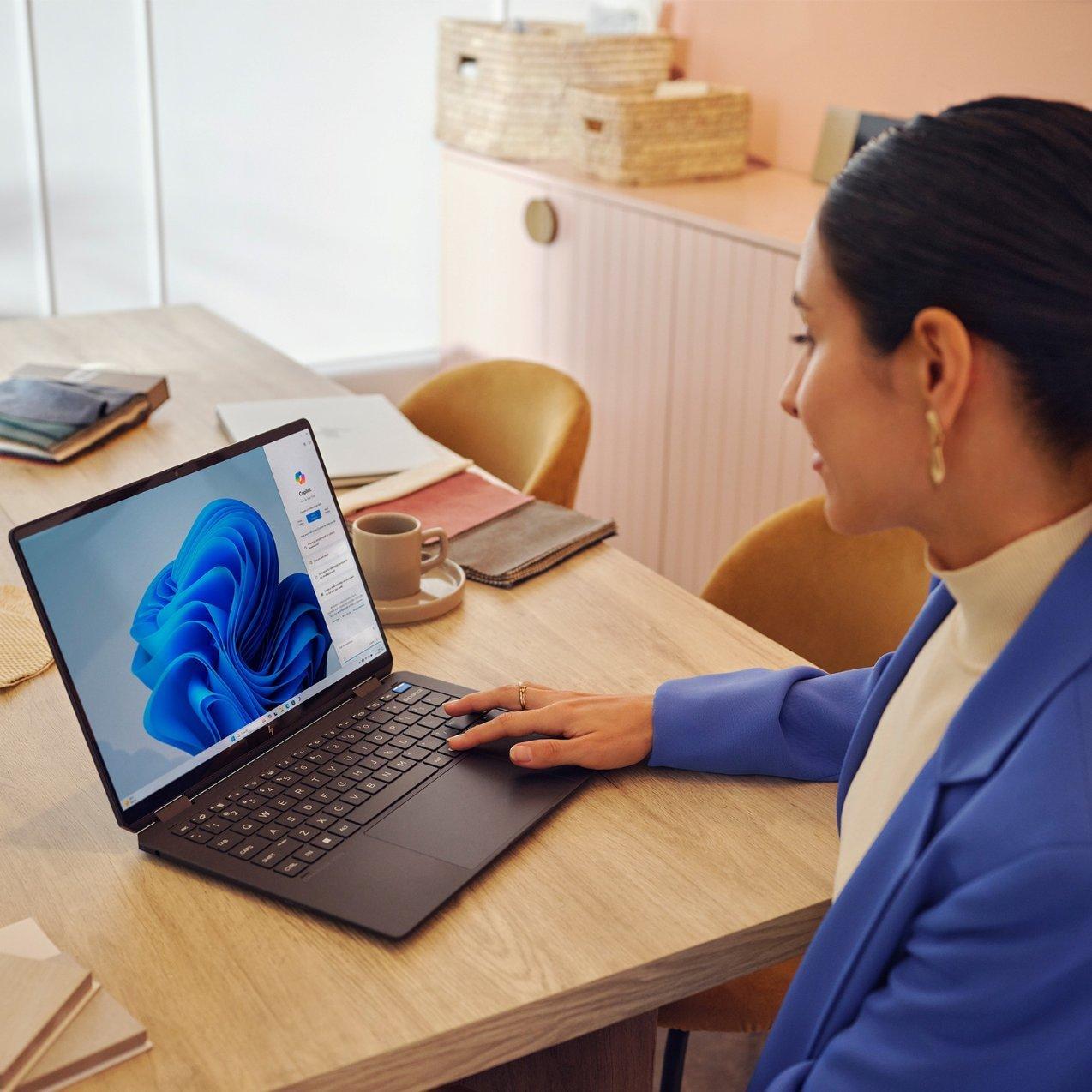A woman in a blue blazer uses an HP laptop on a wooden table. The screen displays Windows 11 with the Microsoft Copilot sidebar open. Fabric samples and a coffee mug rest nearby.