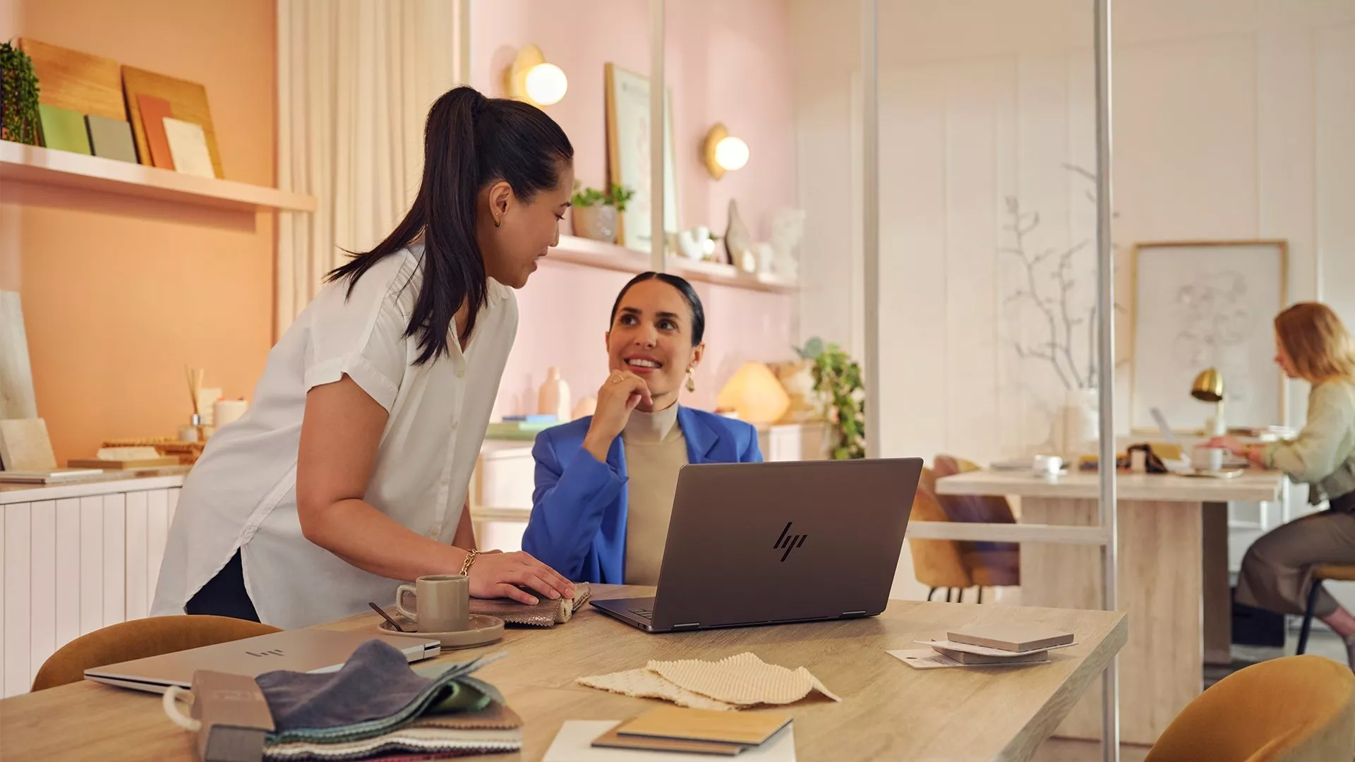 Two women collaborate at a desk with a laptop and fabric samples in a modern office space, smiling and engaged.