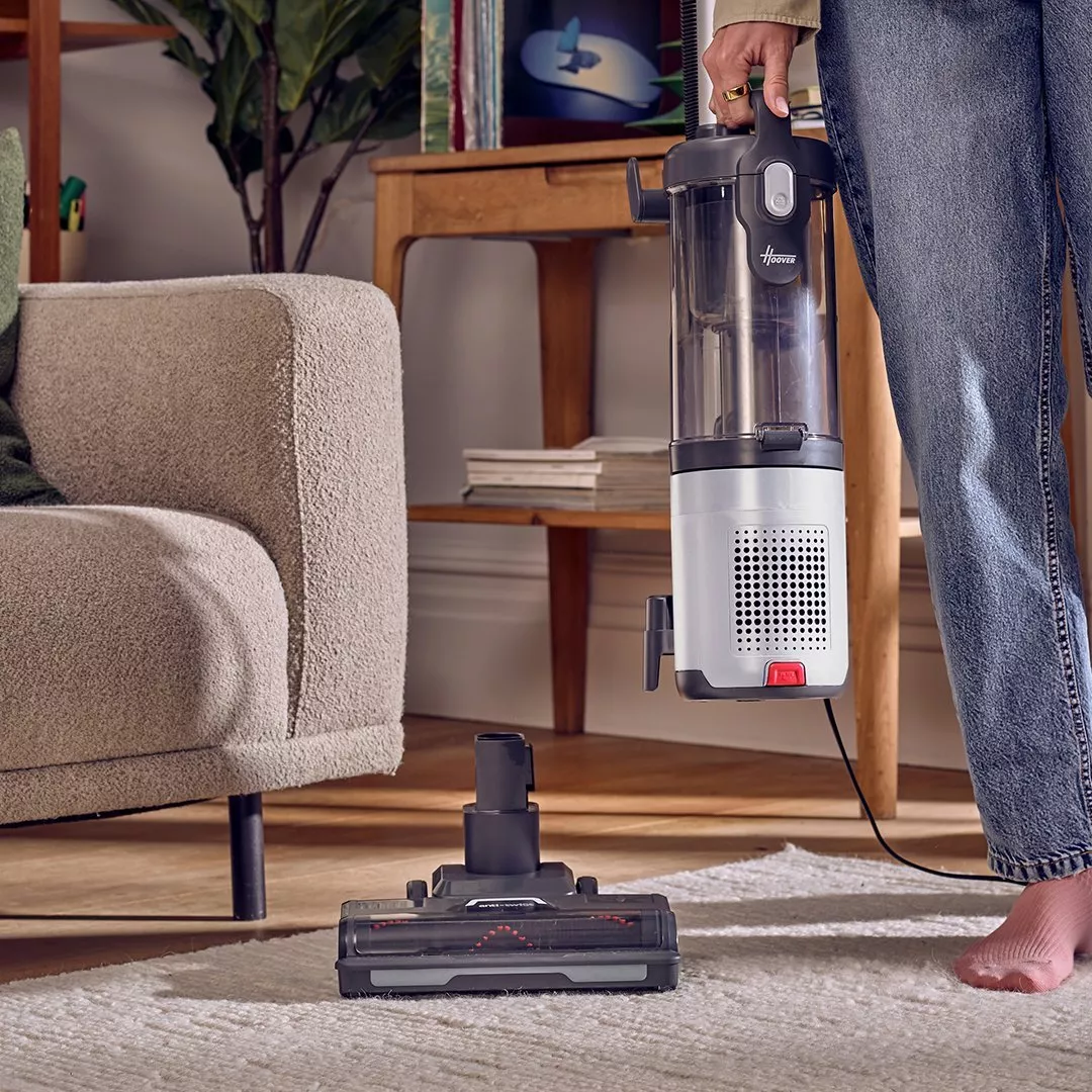 Person holding a Hoover vacuum cleaner near a cosy sofa, with a vacuum head on a carpeted floor.