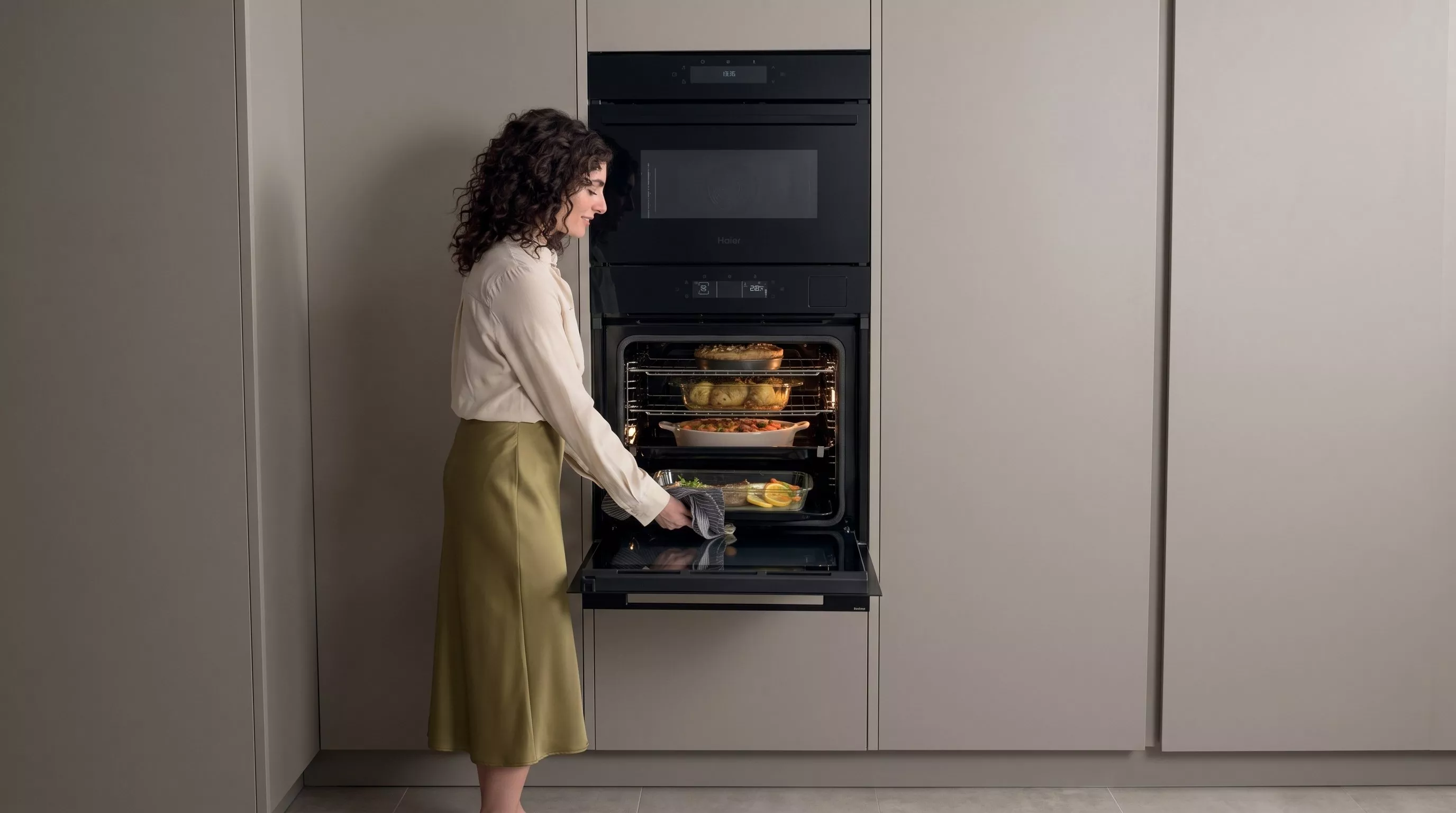 A woman with dark curly hair opens a sleek, built-in black Haier oven while using a striped kitchen towel to check on a glass baking dish with fish and lemon slices.