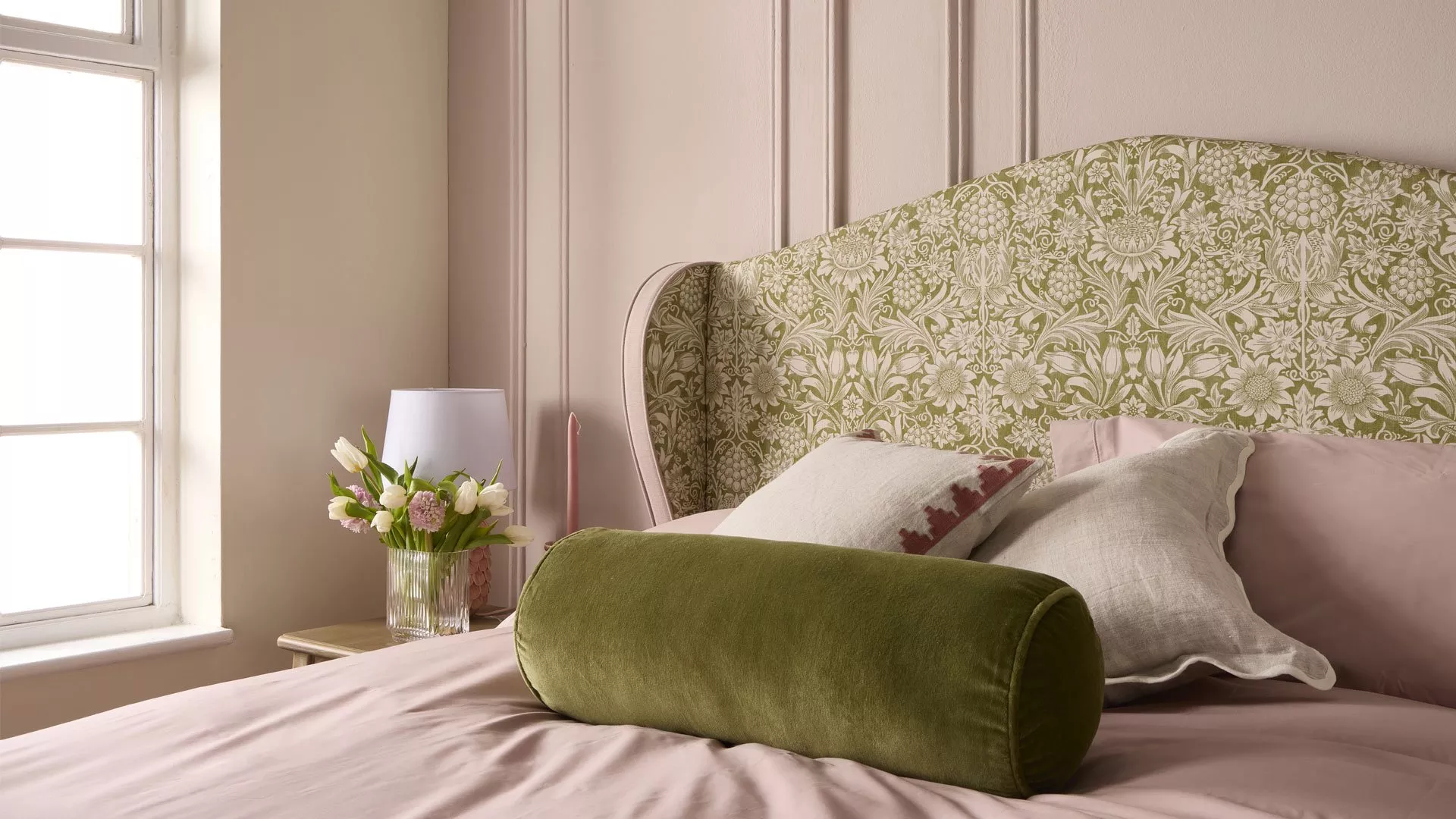 Stylish bedroom with green floral upholstered headboard, pink bedding, velvet bolster cushion and bedside flowers by window