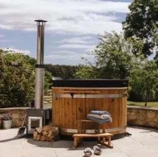 Wood-fired cedar hot tub with chimney on stone patio, towels and sandals, garden and trees behind
