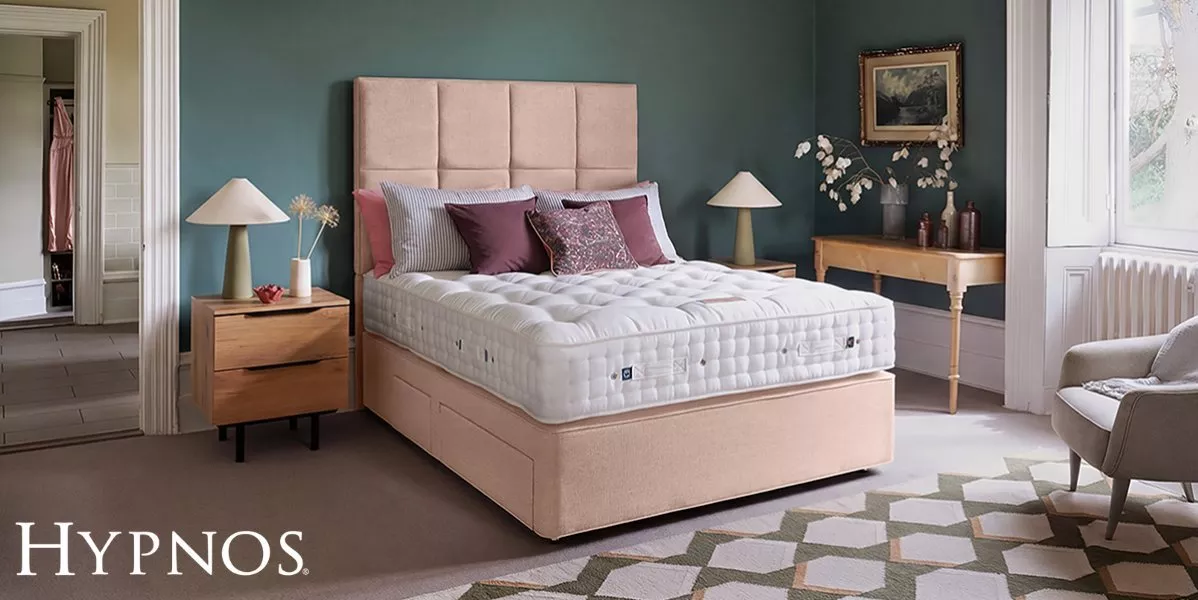 A plush tufted mattress on a light pink upholstered divan bed, styled with plum pillows in a bedroom with teal walls, wooden nightstands, matching table lamps, and a console table.
