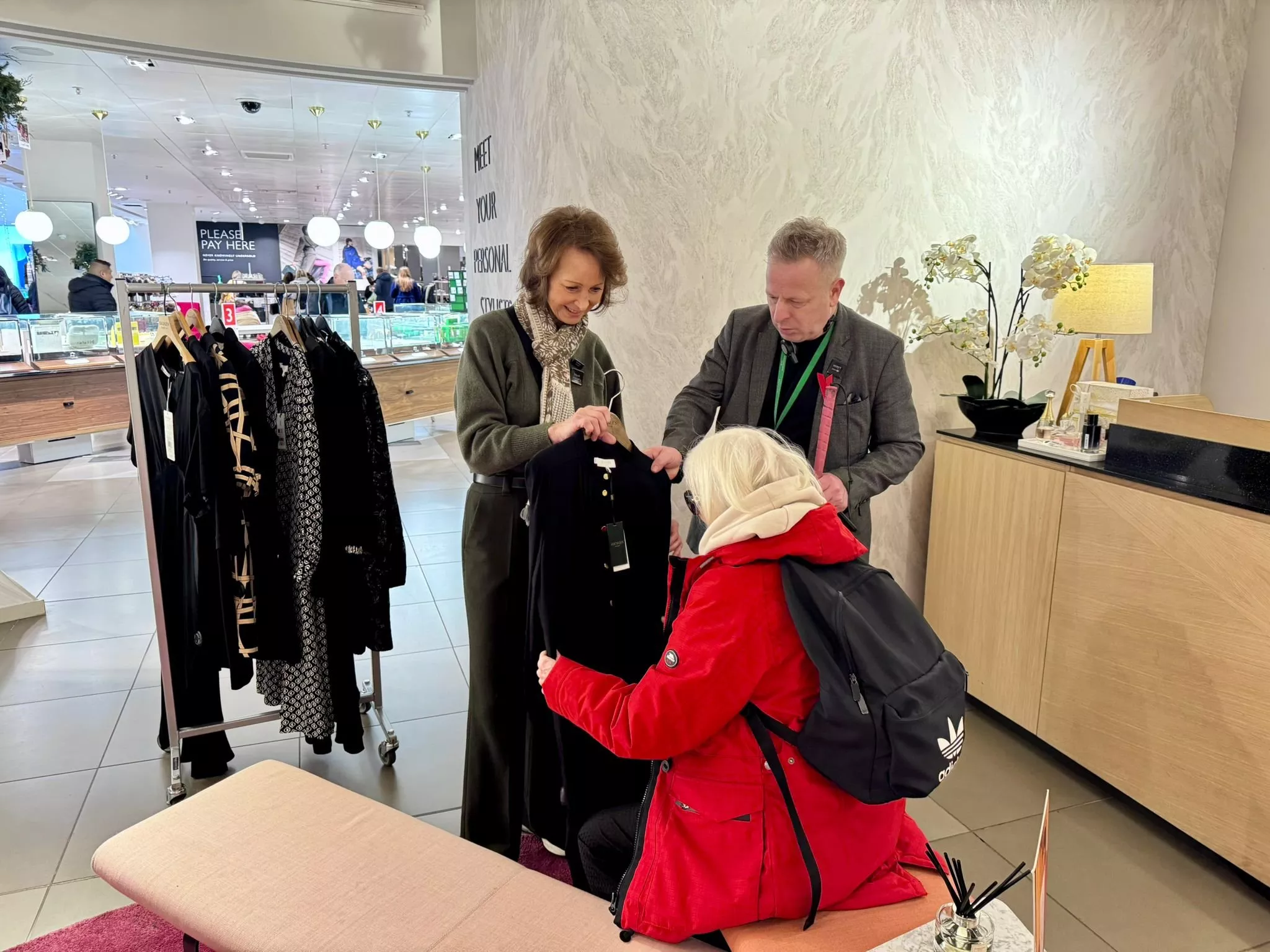 Personal shopping assistants helping a customer choose a black dress in a department store fashion lounge area.