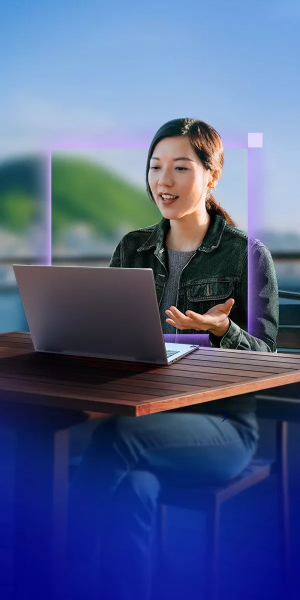 A woman is engaged in a video call on a laptop, sitting outdoors with a scenic blurred background.