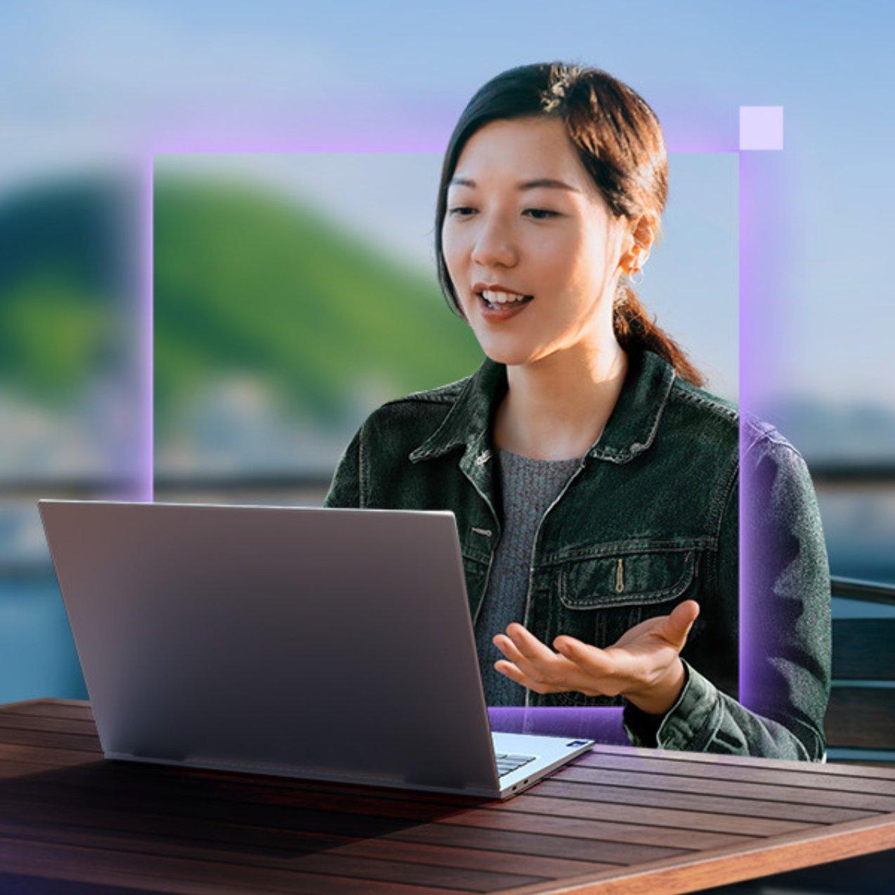 A woman sits at an outdoor table using a laptop, gesturing with her hand while talking during a video call. A glowing purple frame outlines her.
