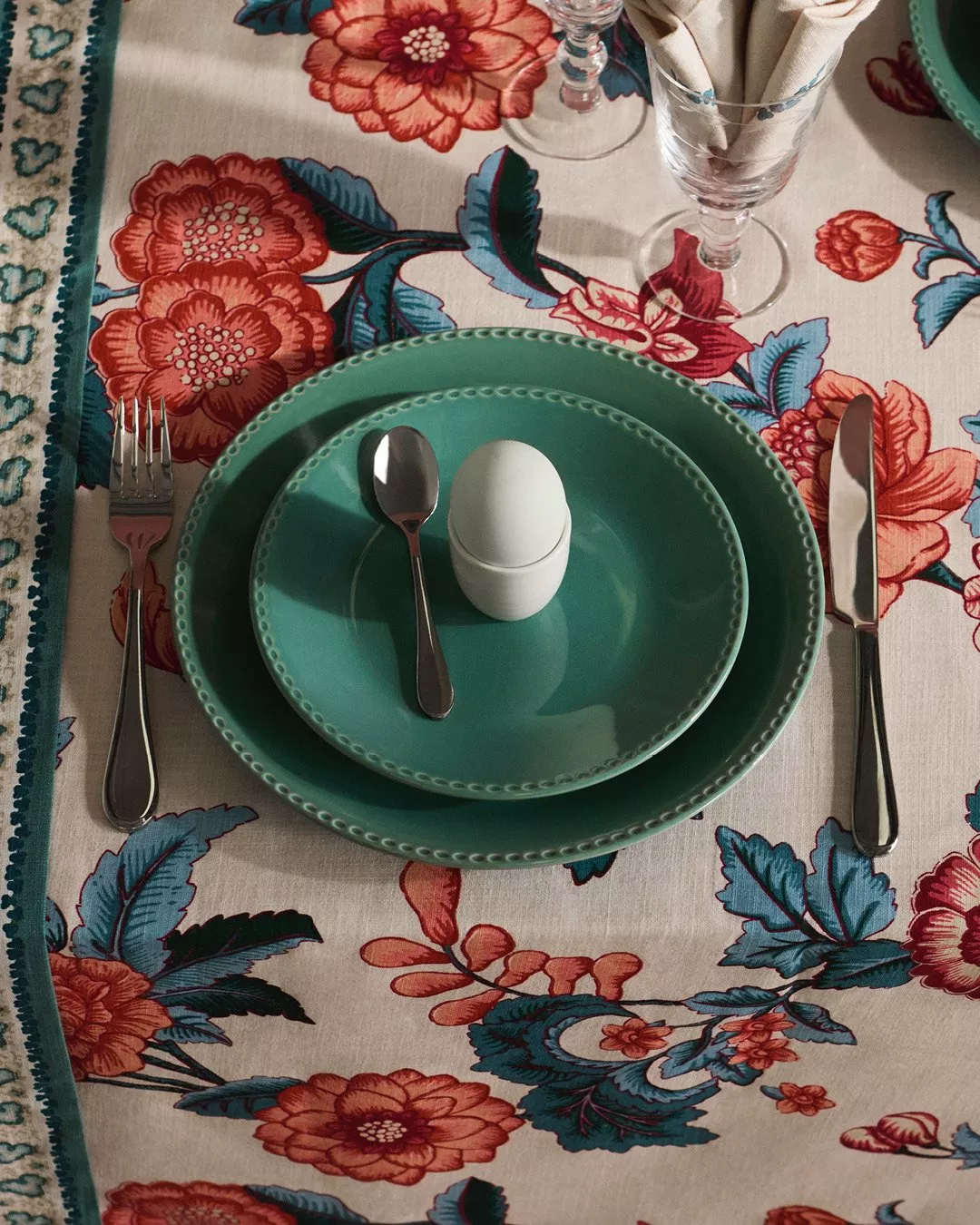 Elegant table setting with teal plates, cutlery, and a white egg-shaped item. Floral tablecloth enhances decor.